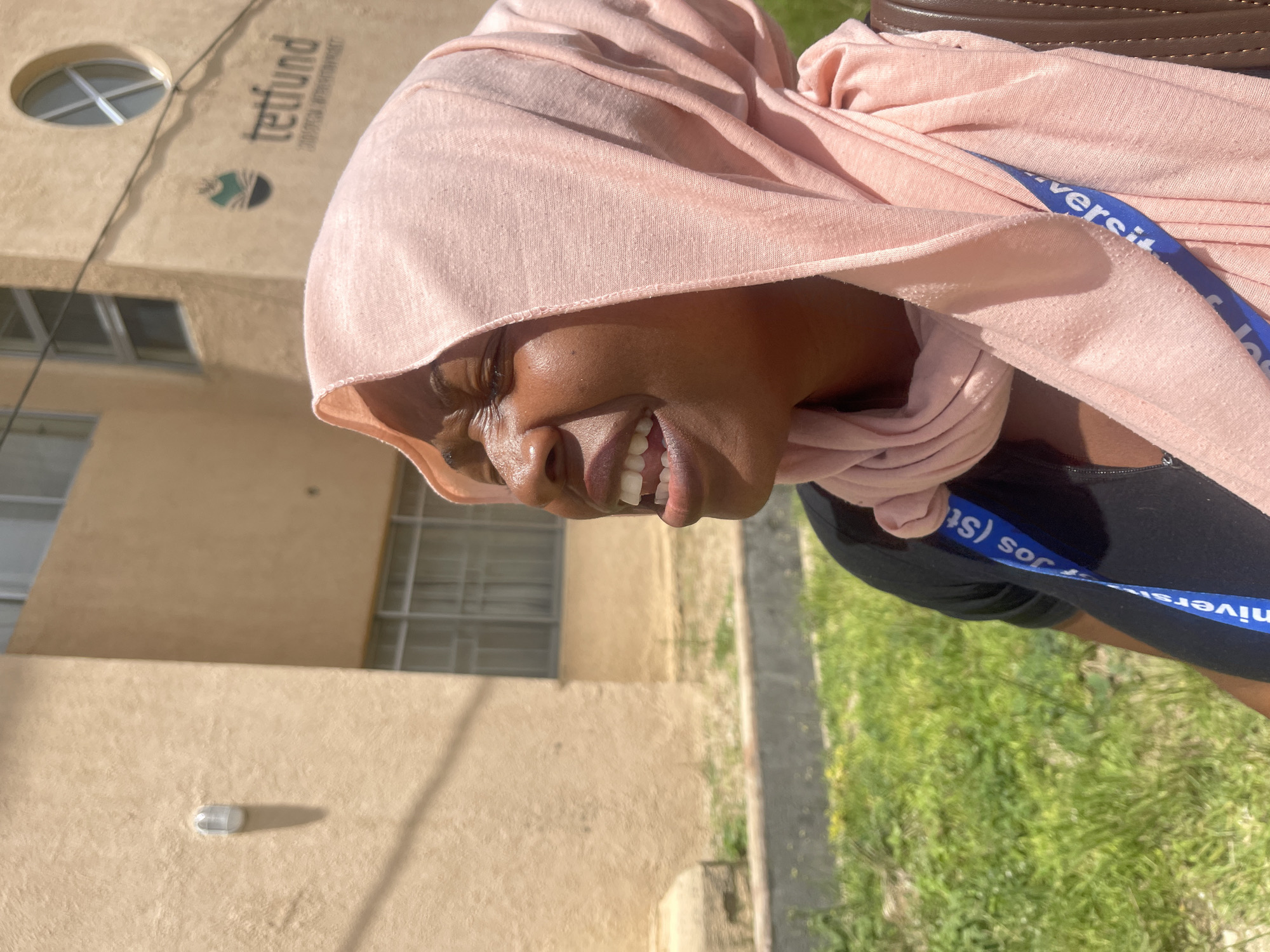 In Rafin Par, Nigeria, a close-up, medium shot captures a woman wearing a light pink headscarf and a black shirt. She is outdoors, with a tan building and green grass in the background. The building features windows, a light fixture, and a logo with the text "tetfund". The woman's face is turned upwards, her eyes are closed, and her mouth is open as if she is speaking or singing. Her skin is shiny, possibly from sweat or oil. A blue lanyard with white text partially visible, reading "Univers Jos (St..." is visible on her shirt. The lighting suggests a sunny day.