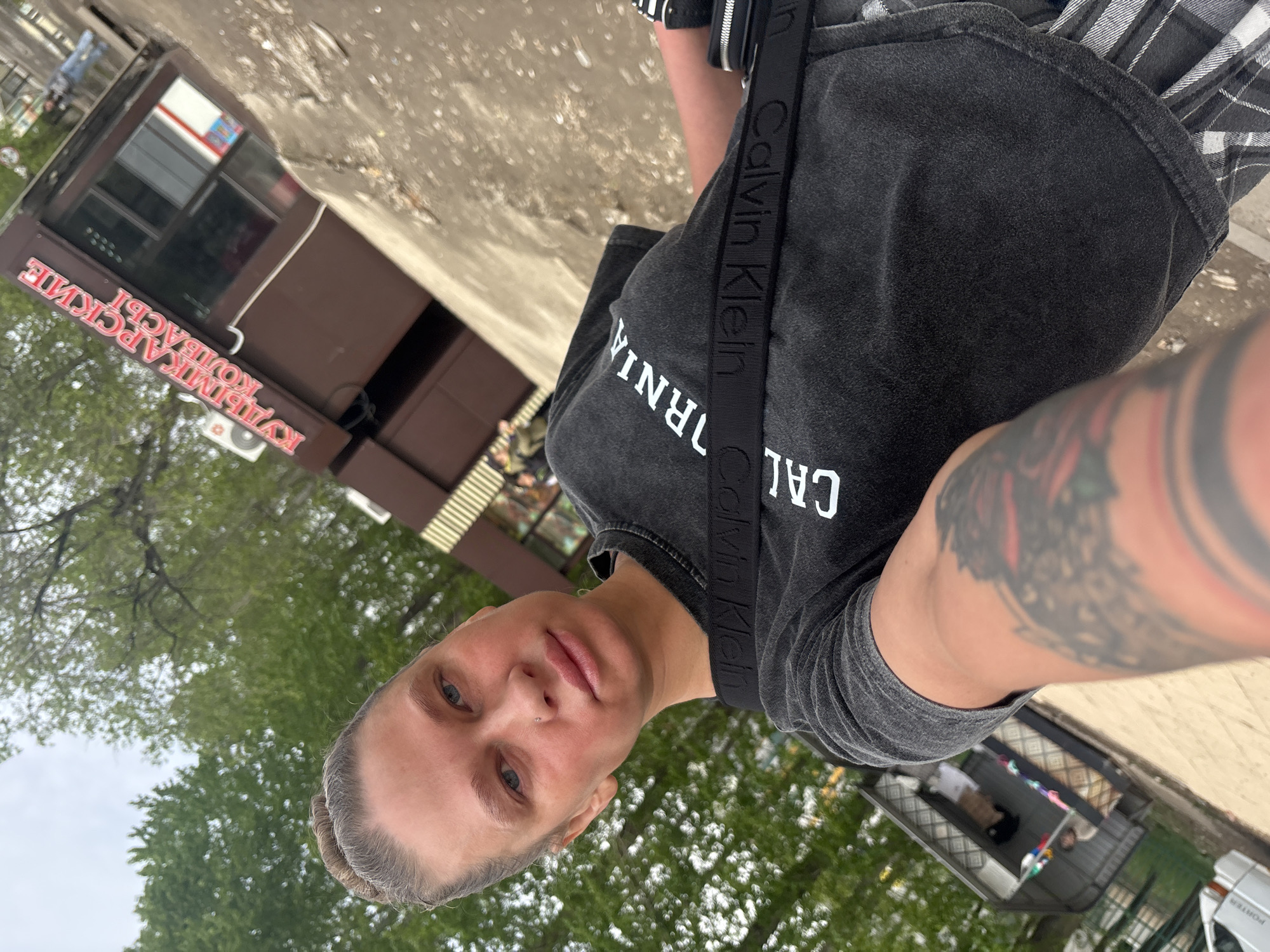 A low-angle photograph shows a woman standing near what appears to be an outdoor food kiosk or shop. The woman has gray hair pulled back, blue eyes, and a nose piercing. She is wearing a gray t-shirt with "California" printed on it and a black Calvin Klein crossbody strap. Her left arm has a colorful tattoo.
The background includes green trees and foliage, suggesting an outdoor setting. The food kiosk has a brown exterior and a sign in Russian: "КУЛЫМКА АРСКИЕ КОЛБАСЫ" (Kulyka Arskiye Kolbasy), which translates to "Kulyma Arsk Sausages." A "PORTER" vehicle is also visible.
The location is identified as Perm, Russia, based on the Russian sign and the overall visual cues.