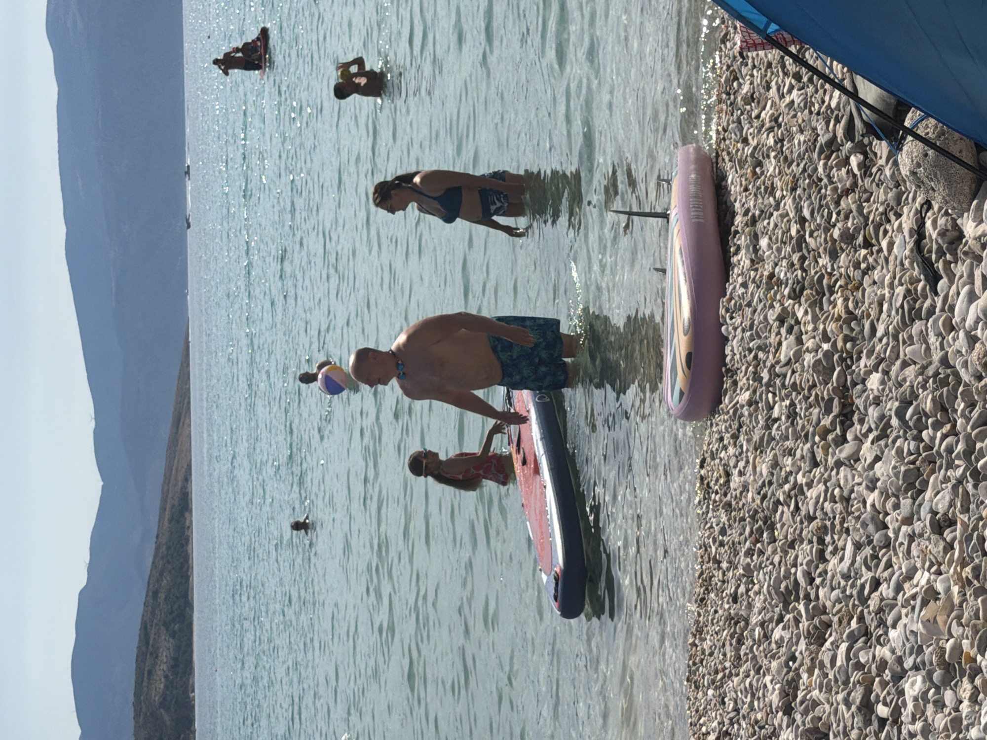 This image depicts a sunny day at a beach in Baška, Croatia. In the foreground, a pebbled shoreline is visible, with a pink inflatable object lying on the stones. The water is clear and shallow, with several people engaged in various water activities. A man in swim trunks stands near a paddleboard, and a young girl in a swimsuit is nearby. Further out in the water, other individuals are visible, some swimming and others on what appear to be boats or inflatable watercraft. In the background, rolling hills and mountains form the backdrop to the coastal scene. The overall impression is one of a recreational beach environment on a warm day.