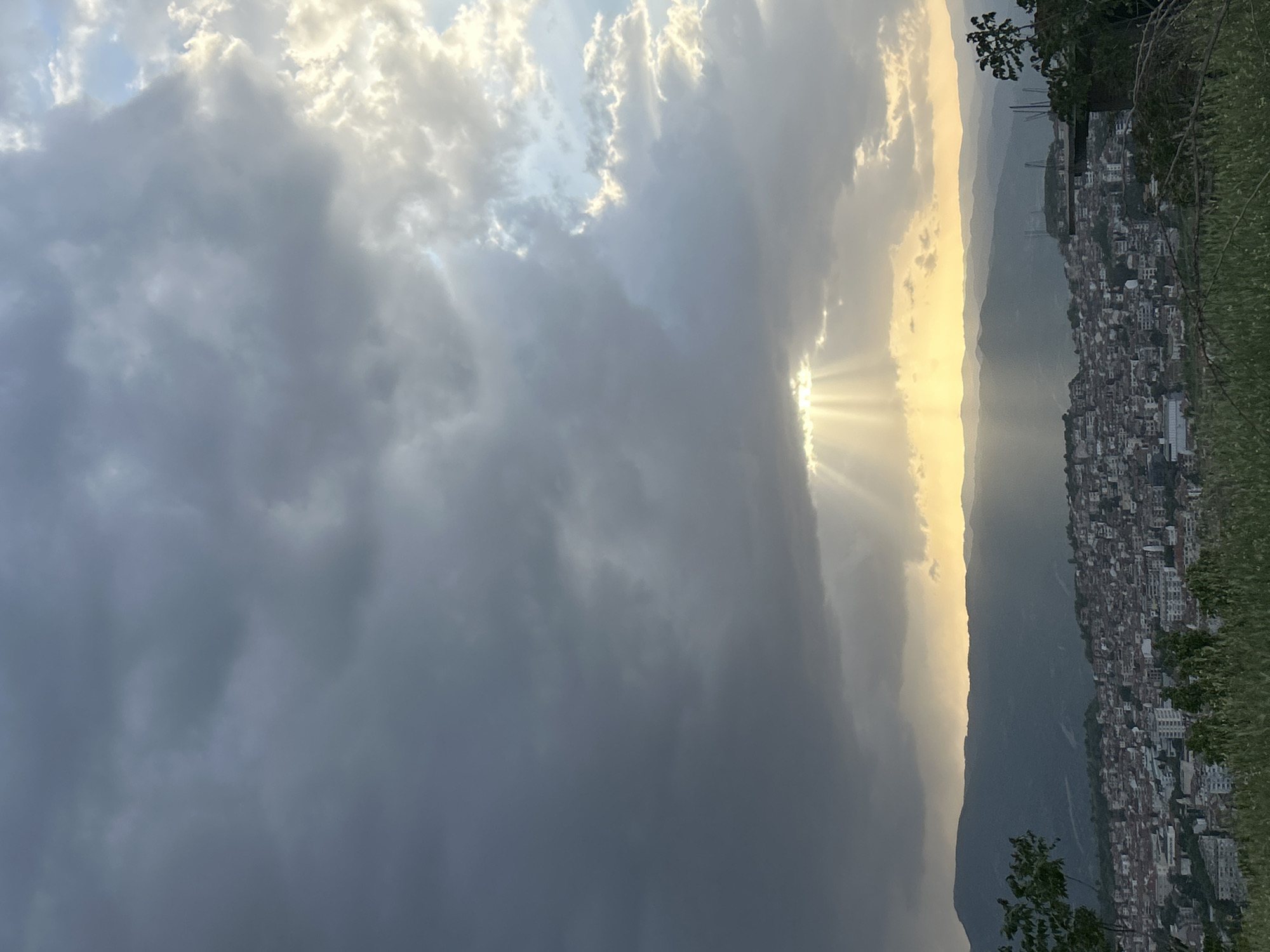 The image shows an outdoor, daytime view of a city nestled on a hillside, likely Polenitsa, Bulgaria, under a dramatic sky. The sky is filled with clouds, ranging from dark, heavy storm clouds on the left to bright, sunlit clouds on the right. Rays of sunlight break through a gap in the clouds. A series of mountains are visible in the background. The city consists of closely packed buildings that extend up the slope. Trees are at the top right corner of the image.