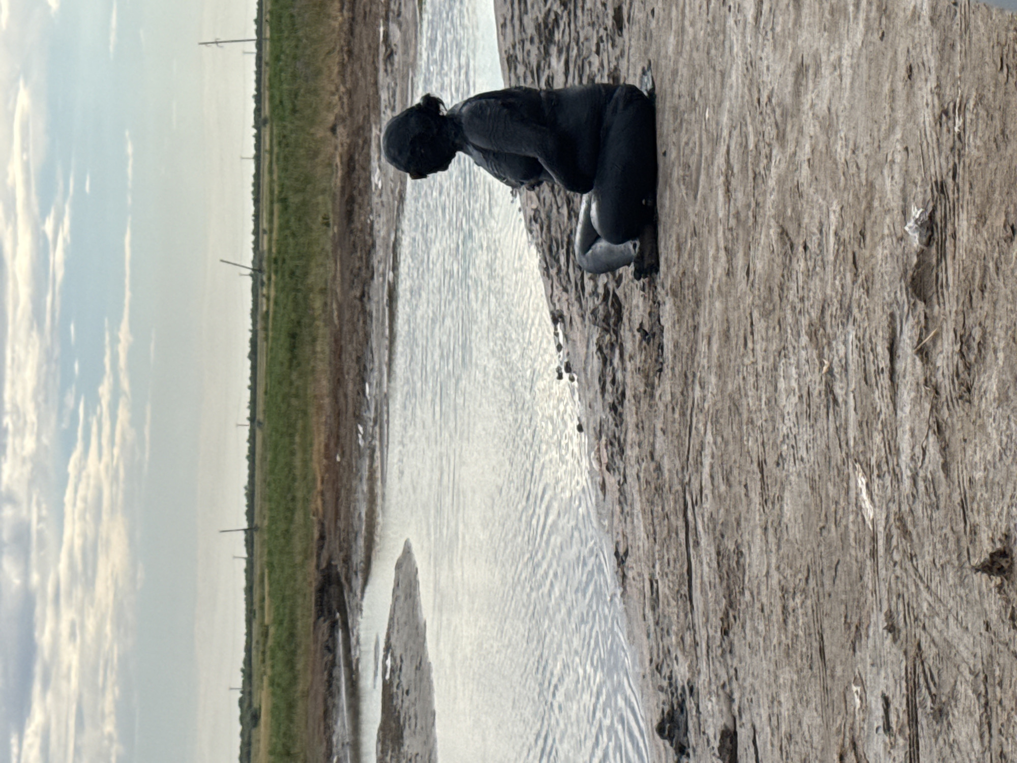 A mud-covered person sits cross-legged on the muddy shore of a body of water. The person is facing to the right, with their head slightly bowed and their arms resting on their lap. The skin of the person is entirely covered in dark mud, obscuring any distinguishing features except for their silhouette and the shape of their body. Their hair appears to be pulled back, also covered in mud, with what look like dark goggles or eye coverings visible. The water in front of them is calm with small ripples. The surrounding environment is a barren, muddy landscape with sparse vegetation in the distance. The ground is cracked and dry in places. In the far background, a flat, green plain is visible under a cloudy sky with thin, horizontal clouds. There are a few distant, thin poles or structures dotted across the landscape. The location is identified as a natural mud pool in the Yamyshevskoye rural district, Kazakhstan.