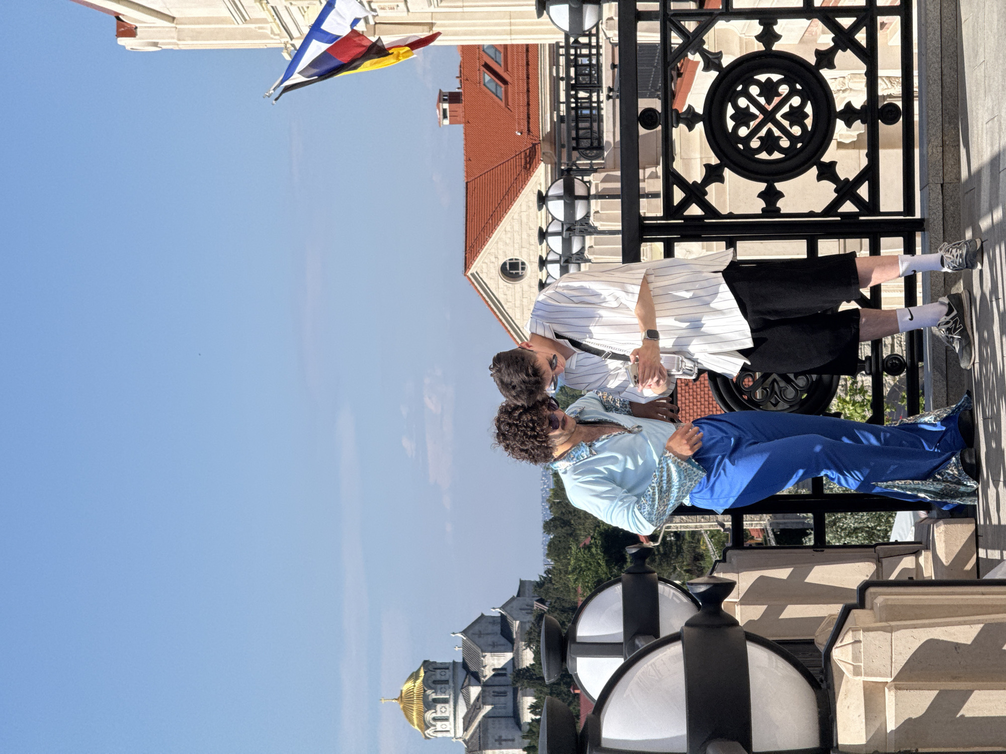 In the city of Unknown, Unknown, on a sunny day, two young men stand on a balcony, facing each other and looking down at a smartphone held by the man on the right. The man on the left wears a sparkly blue shirt and bright blue flared pants. The man on the right is dressed in a white and blue striped short-sleeved shirt, black shorts, white socks, and grey sneakers. Behind them, a building with a red roof and a neoclassical facade is visible, with flags flying from its upper levels. In the distance, a large building with a golden dome, resembling a church or cathedral, is situated amongst green trees. The balcony features a black metal railing with decorative elements and two large, cylindrical lamps.