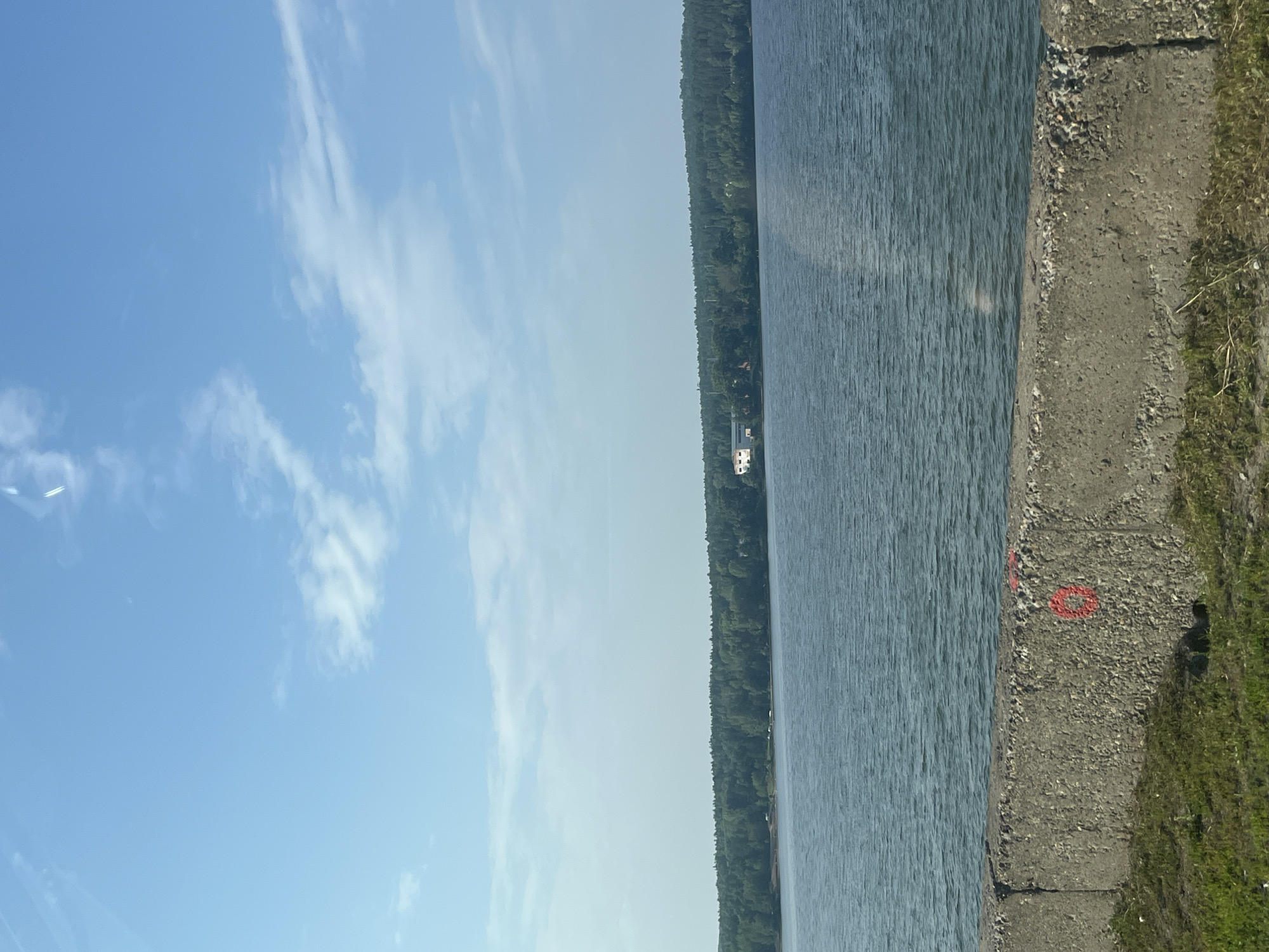 The image shows a wide, calm body of water, likely a reservoir or river, under a clear blue sky with sparse white clouds. The water stretches to a distant, tree-covered shoreline. On the far shore, amidst the dense green foliage, a white building with a light-colored roof is visible, suggesting a residential or industrial structure. In the foreground, a concrete barrier, possibly a retaining wall or embankment, runs horizontally across the lower portion of the frame. This barrier is textured and shows signs of weathering. Patches of grass grow in front of and behind the barrier. There are some red spray-painted markings on the concrete barrier, including what appears to be a circle. The overall scene depicts a tranquil natural environment adjacent to human structures. This location is likely along the Kama River in the city of Chaikovsky, Russia, given the presence of the wide river and the visual characteristics that align with the area.