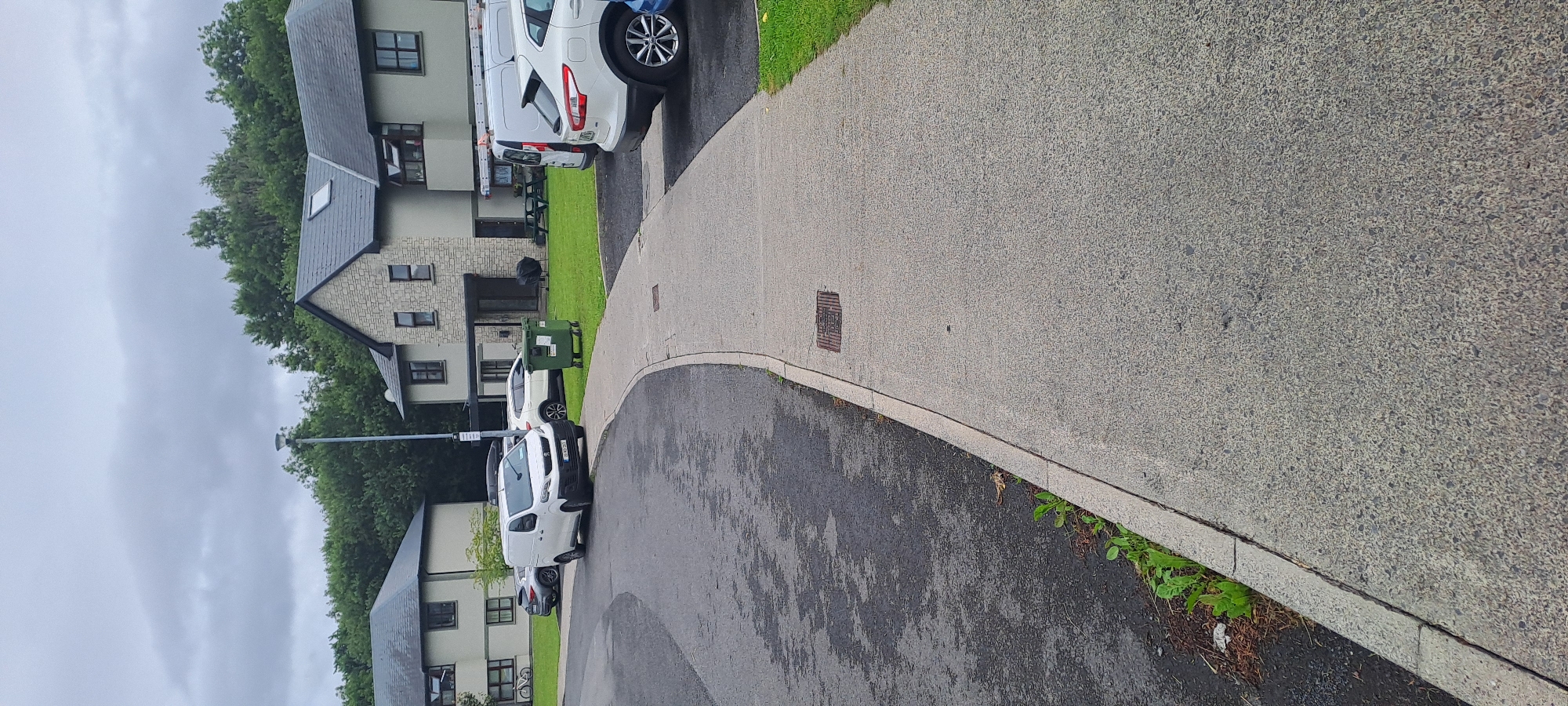 The image depicts a residential area in Castlebar, Ireland. A paved walkway curves from the foreground towards a row of modern townhouses, some with stone facade accents. Several vehicles are parked along the walkway and on the street, including a white panel van, a white SUV, and a dark sedan. A green recycling bin is positioned on the grass in front of one of the buildings. Trees and greenery surround the residential buildings, and the sky is overcast. The scene appears calm and ordinary, with no visible human activity.