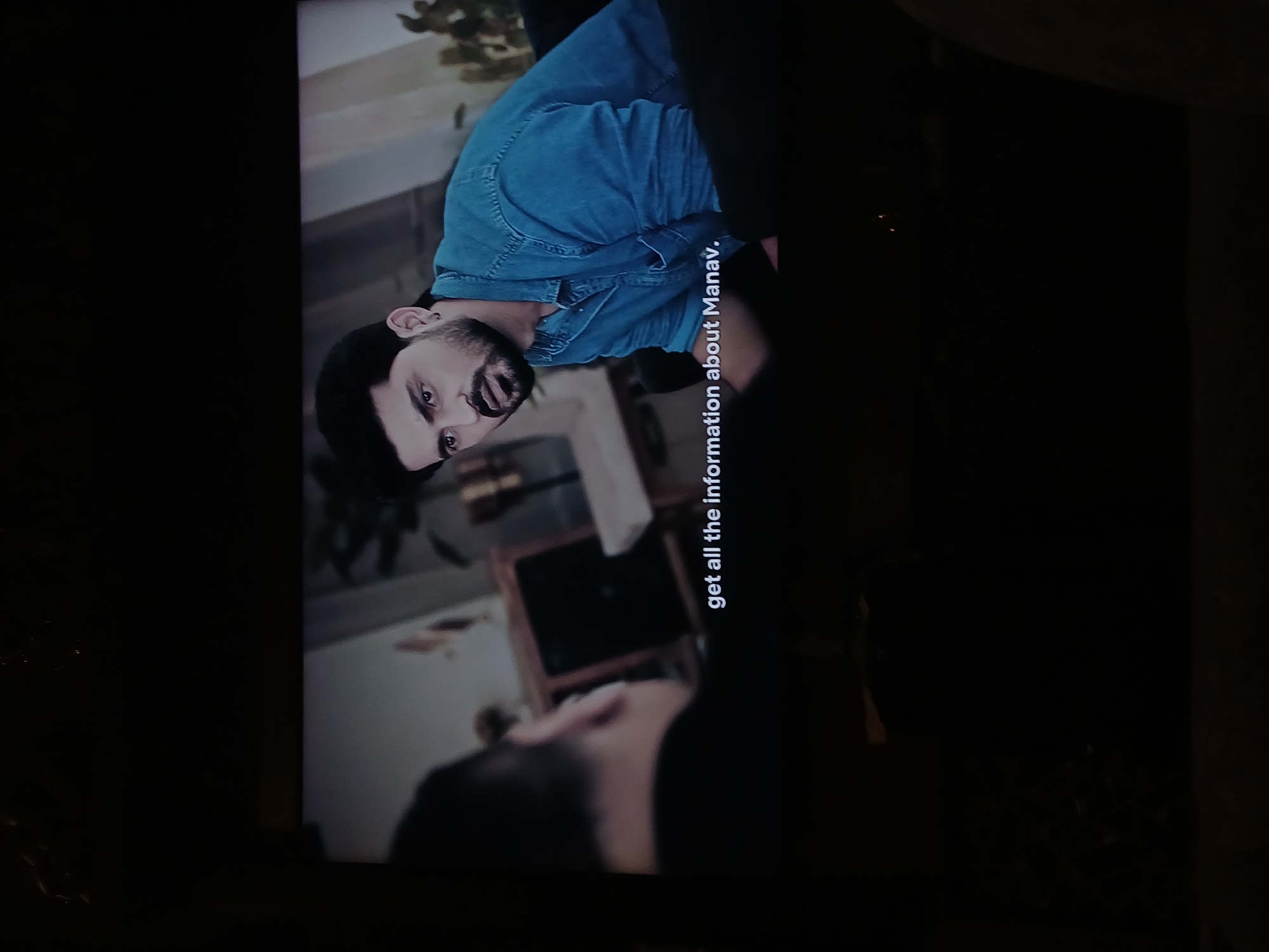 Here's a description of the image:
A person is looking down at another person. The first person has dark hair and a short beard, and is wearing a denim shirt. The second person's face is not clearly visible, but they appear to have dark hair. Text on the screen says "get all the information about Manav." The setting appears to be indoors, possibly a living room, with glimpses of furniture and decor. There are no indications that show the location is in Bengaluru, India.