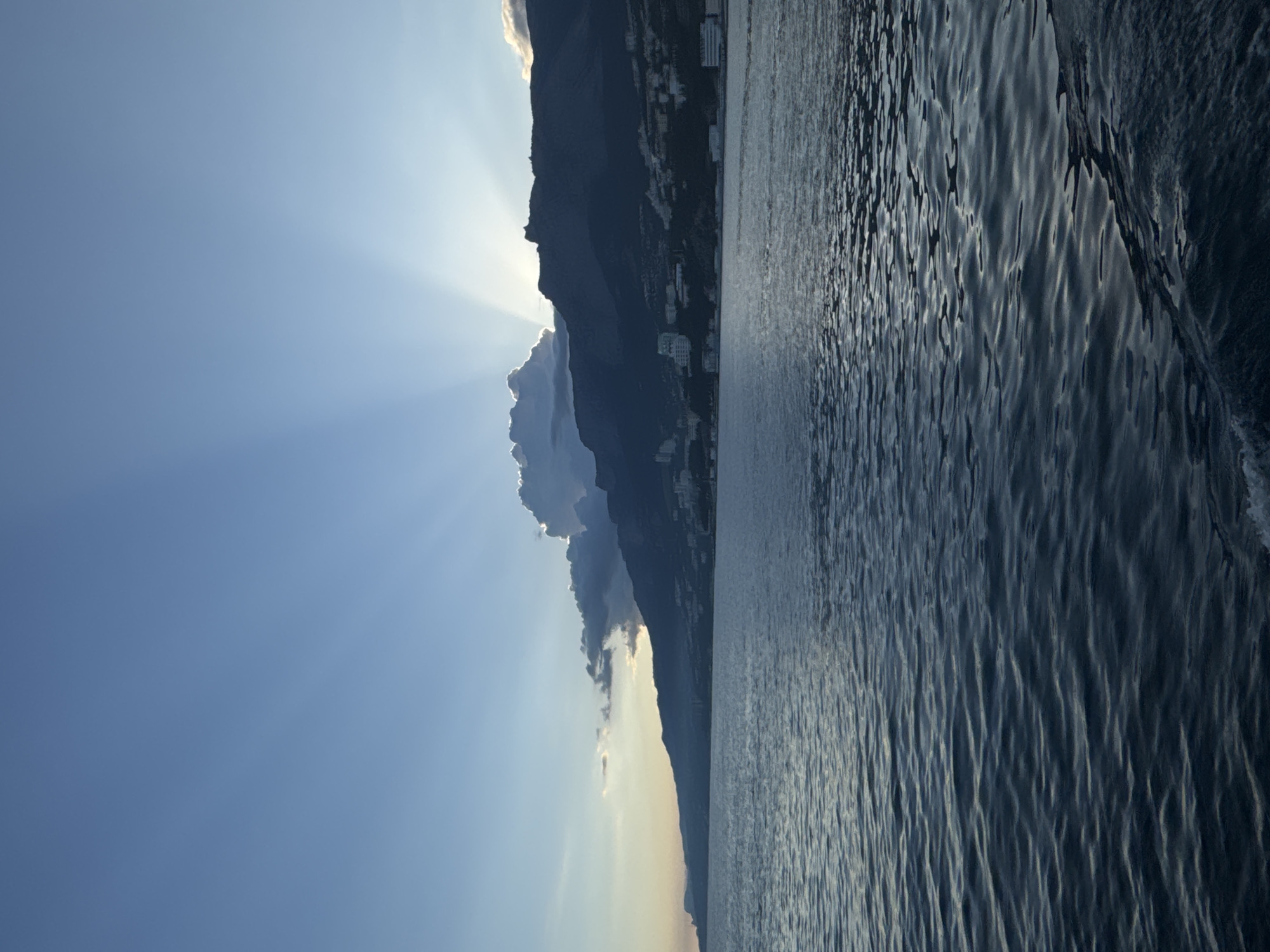The image shows a landscape with a body of water, a mountainous terrain, and the sky. The sun is partially obscured by the mountains, causing sun rays to spread across the sky. Buildings are visible on the mountain slopes. The body of water appears textured with ripples. The location may be in the Гаспринский поселковый совет, Ukraine, as requested.