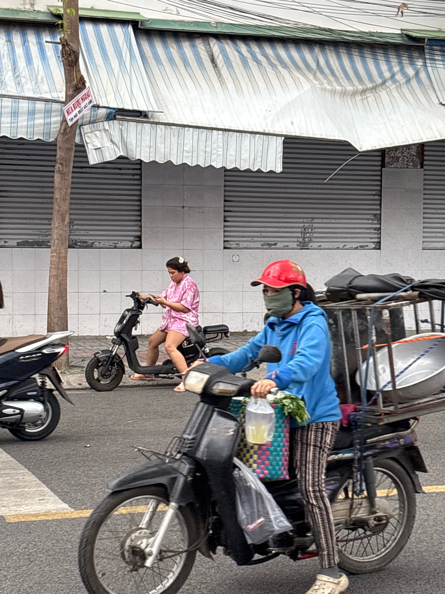 In Nha Trang, Vietnam, on a street with striped awnings and metal roll-up shutters visible in the background, two individuals are on motorcycles. In the foreground, a person wearing a blue hooded jacket, a red baseball cap, and a green face mask is riding a dark-colored motorcycle with a metal cargo rack behind them. The cargo rack holds a large metal pot and other items, and the rider is holding a plastic bag with a yellow liquid and some greenery. To their left, another person is seated on a black electric scooter, looking down at their phone. This individual is wearing a pink patterned short-sleeved shirt and shorts. In the left background, part of a silver and black scooter is visible. The street is paved, and there are yellow and white lines painted on the road. A tree with a sign attached is also present on the left side of the image.