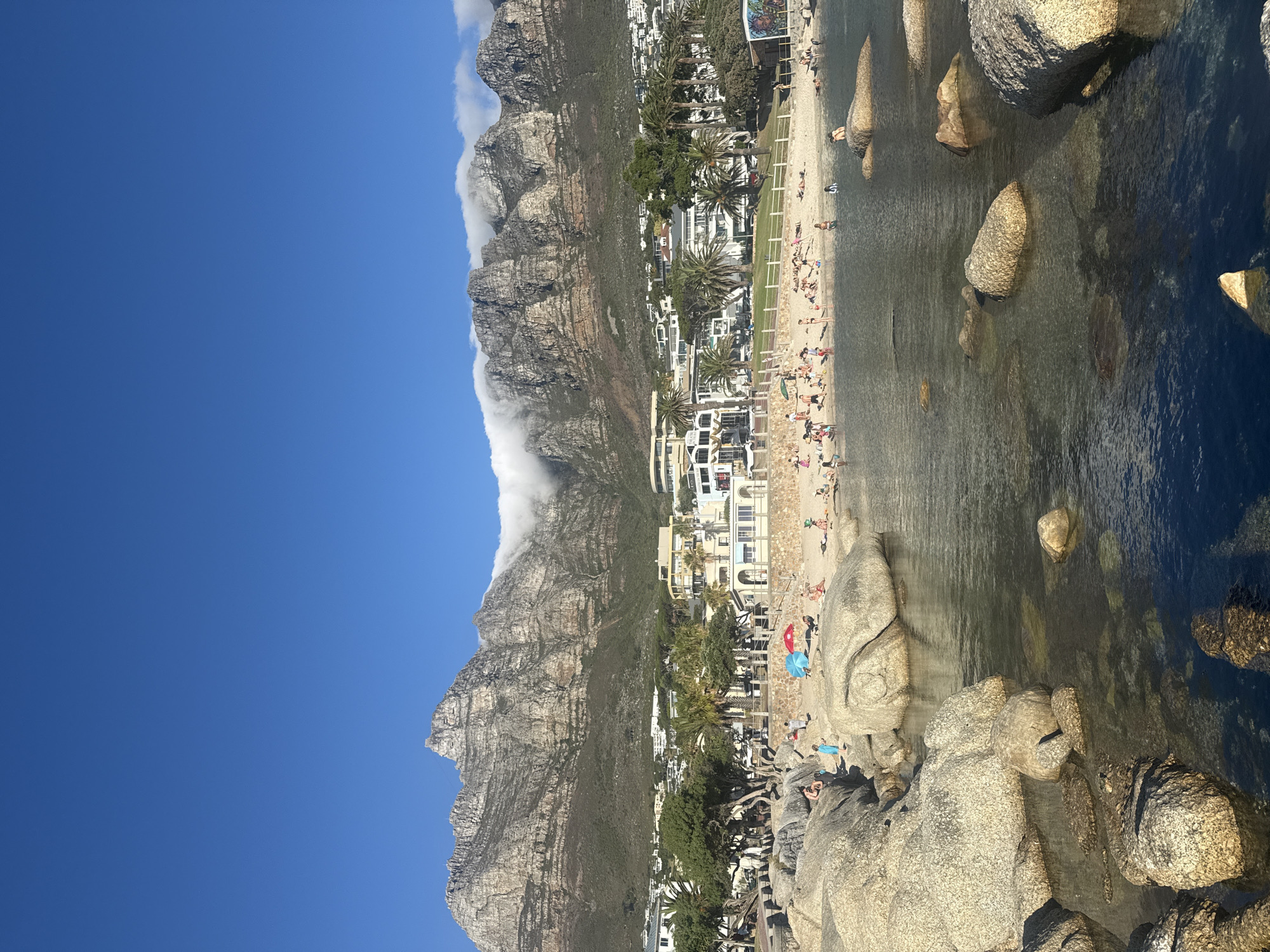 The image shows a landscape in Cape Town, South Africa. A large mountain dominates the scene, with a band of clouds partially covering its peak. The mountain slopes down to a beach area where people are present, either swimming or relaxing on the sand. Buildings are visible along the beach behind palm trees. Large boulders are situated in the water near the shore, and the ocean extends into the distance. A bright blue sky fills the upper portion of the image.