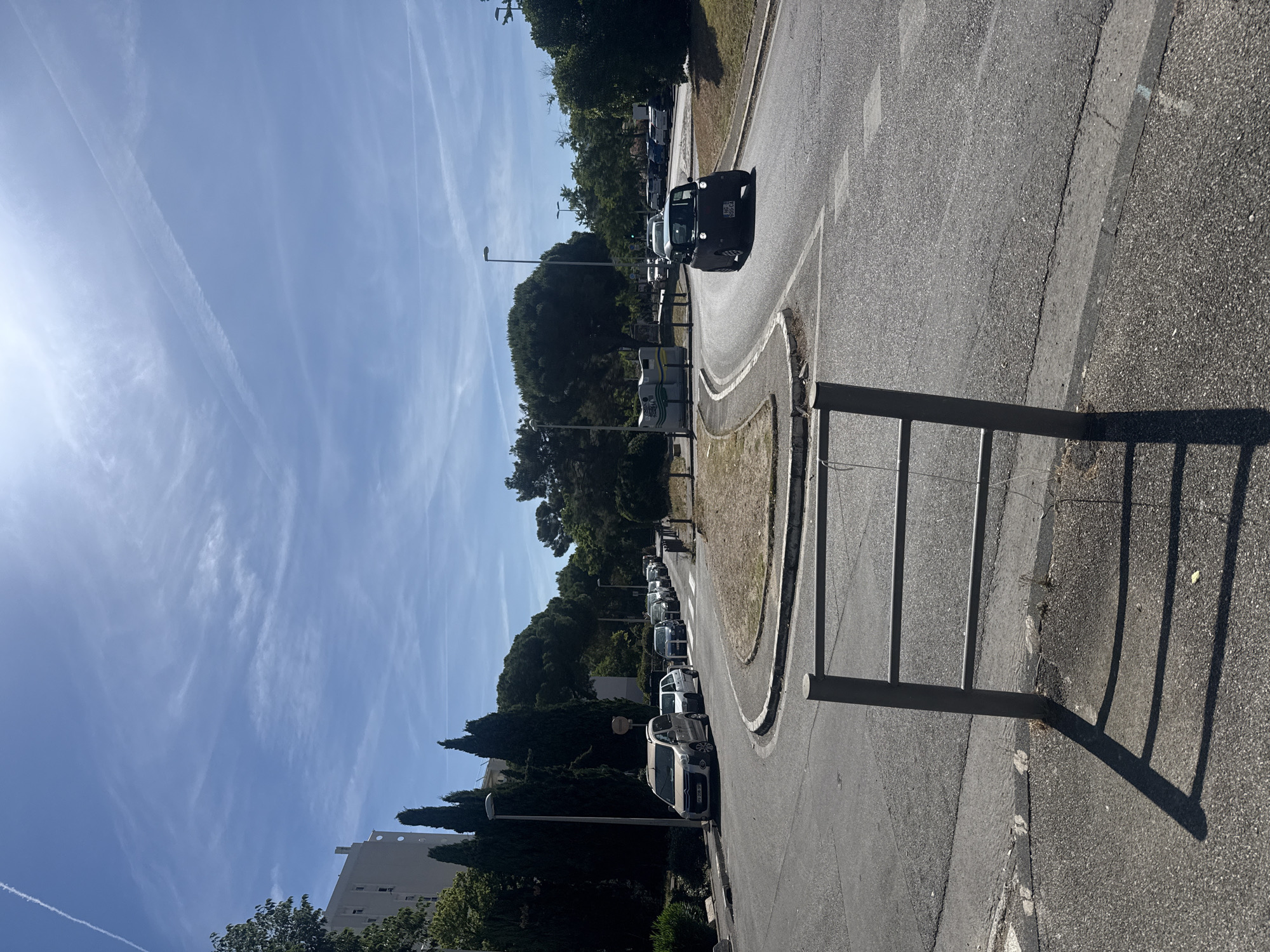 The image shows a road with a curve in a city environment under a bright, partly cloudy sky with contrails from airplanes. In the foreground, a metal railing with three horizontal bars casts a shadow on the asphalt. The road is paved and shows some wear and tear.
On the left side of the road, a row of parked cars is visible, including a light-colored van and several other vehicles. Behind the parked cars, there are tall cypress trees and a building.
On the right side of the road, a dark-colored, small car is driving around the curve. Further down the road, more trees and some buildings are visible. There are streetlights present along the road. The scene appears to be an urban or suburban area, possibly a residential or commercial district, given the presence of buildings, parked cars, and roads. The overall impression is of a sunny day in a developed area.