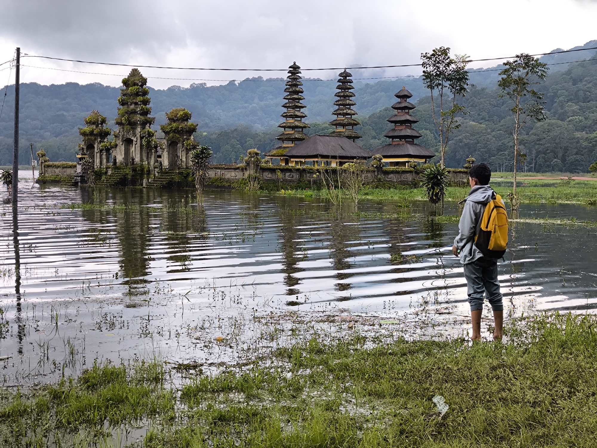 A man with a yellow backpack stands in the water near a green grassy bank, facing a complex of weathered stone structures with tiered roofs, reflecting in the water. The structures are likely temples or shrines, adorned with moss and vegetation, connected by a stone wall. Trees stand to the right of the temples, and a forested hill rises in the background under an overcast sky. Power lines stretch across the top of the frame. The location is in the city of Unknown, Indonesia. The man appears to be observing the site.