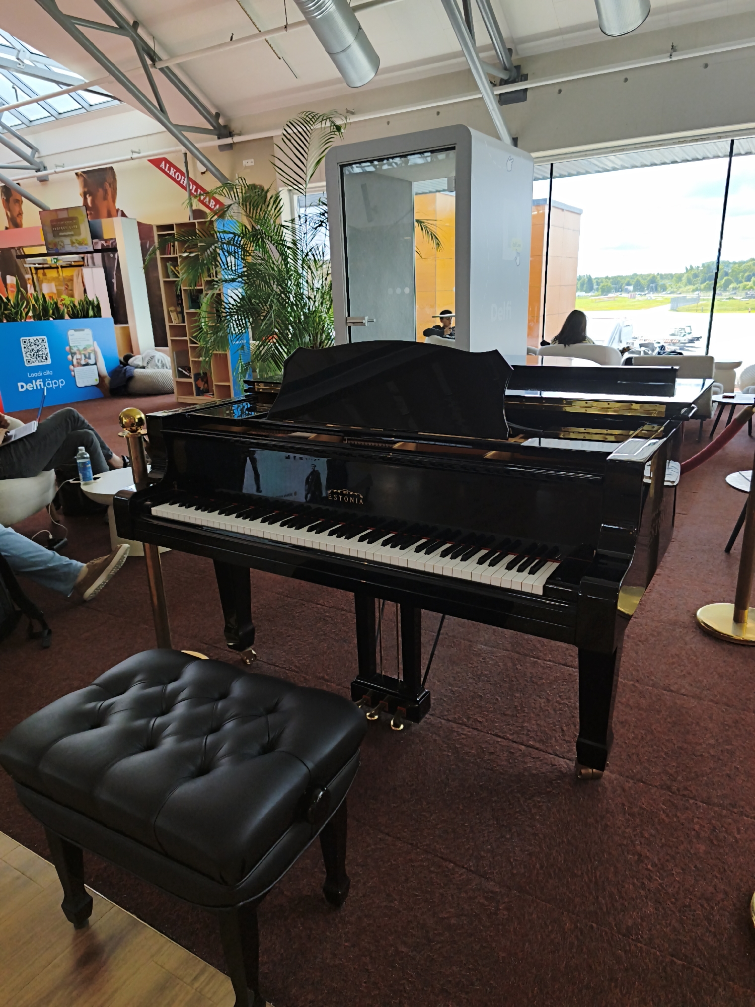 This is a medium shot, eye-level photograph taken indoors at Tallinn Airport in Tallinn, Estonia. The primary subject is a black grand piano with a black piano bench in front of it. The piano is positioned on a reddish-brown carpet. The background includes a large window showing an airfield and a cloudy sky. To the left of the piano, there are people seated in chairs, a bookshelf, and large posters with images and text. There is also a large potted palm tree to the left of the piano and a modern white structure to the right. The lighting appears to be natural light coming from the window and artificial overhead lighting.