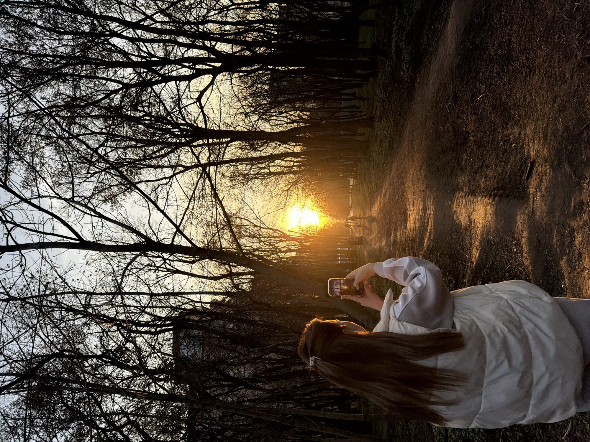 The scene depicts a park pathway in Rostov-on-Don, Russia, at sunset. A female individual, observed from the rear, occupies the left-center foreground. She has long, dark brown hair, partially secured with a light-colored clip, and wears a light-toned puffer vest over a bicolored long-sleeved garment (light grey body, white sleeves) and light-colored pants. She is actively holding a smartphone horizontally with both hands, its screen reflecting the bright light source ahead, consistent with capturing an image or video. The primary light source, a bright orange-yellow sun, is positioned low on the horizon, illuminating a dirt path that extends centrally into the background. Both sides of the path are densely bordered by bare deciduous trees, silhouetted against the gradient sky, which transitions from light blue overhead to bright orange-yellow near the sun. Multiple indistinct human figures, heavily silhouetted, are visible walking further down the sunlit path. A dark, multi-story building with discernible window lines is partially visible through the upper tree canopy on the left side of the frame.