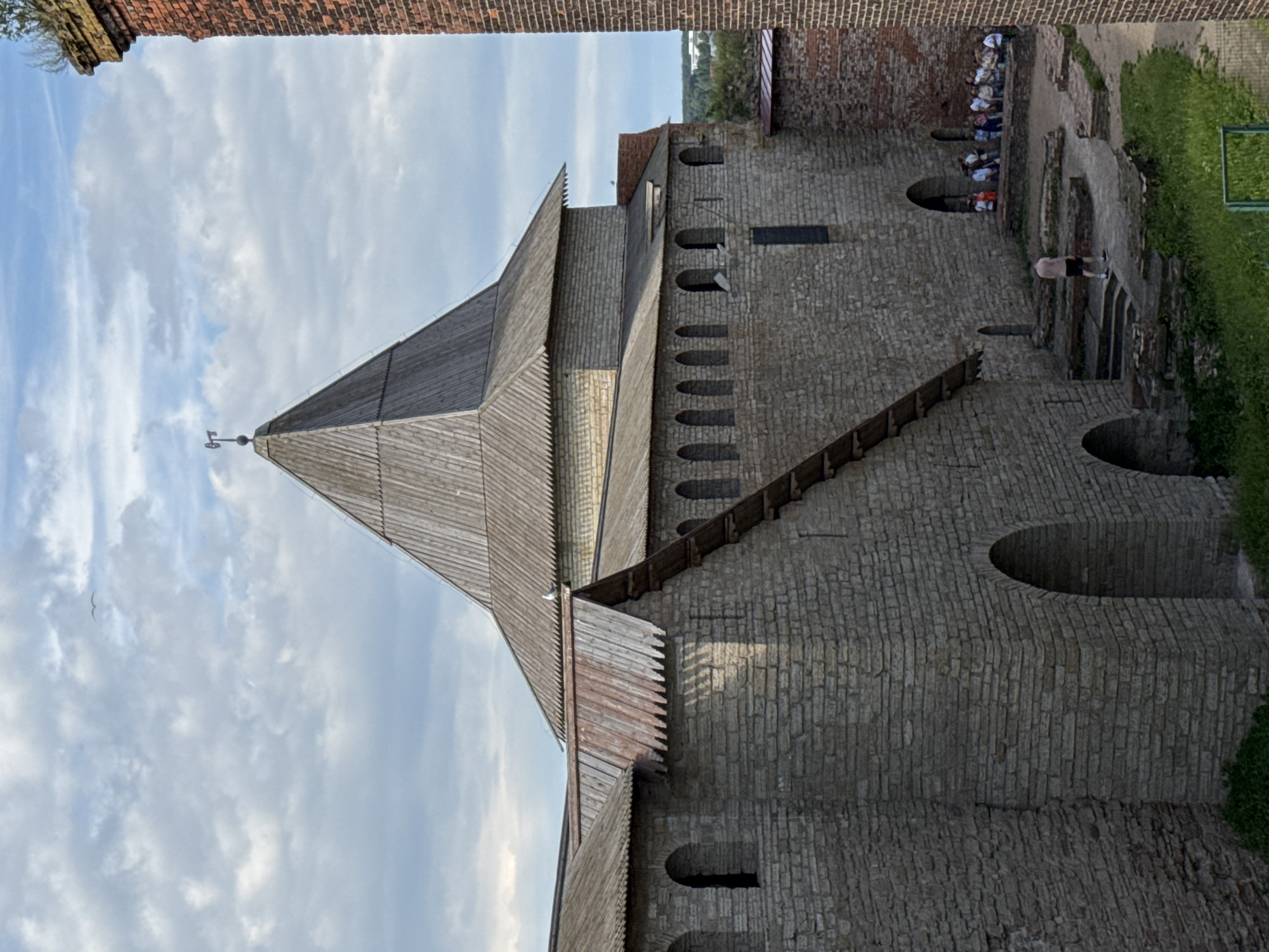 This is an outdoor, eye-level, vertical, full shot of an ancient stone fortress with a wooden spire and roof. The fortress is made of grey stone blocks and has several arched windows and openings. A wooden staircase ascends along the side of the fortress. A few people are visible in the background, some standing and some sitting. One person is walking on a path. The sky is partly cloudy. The sun appears to be setting, casting shadows on the fortress walls. The scene is calm and peaceful. The location is likely Shlisselburg, Russia, given the distinctive architecture. There is no visible text in the image.