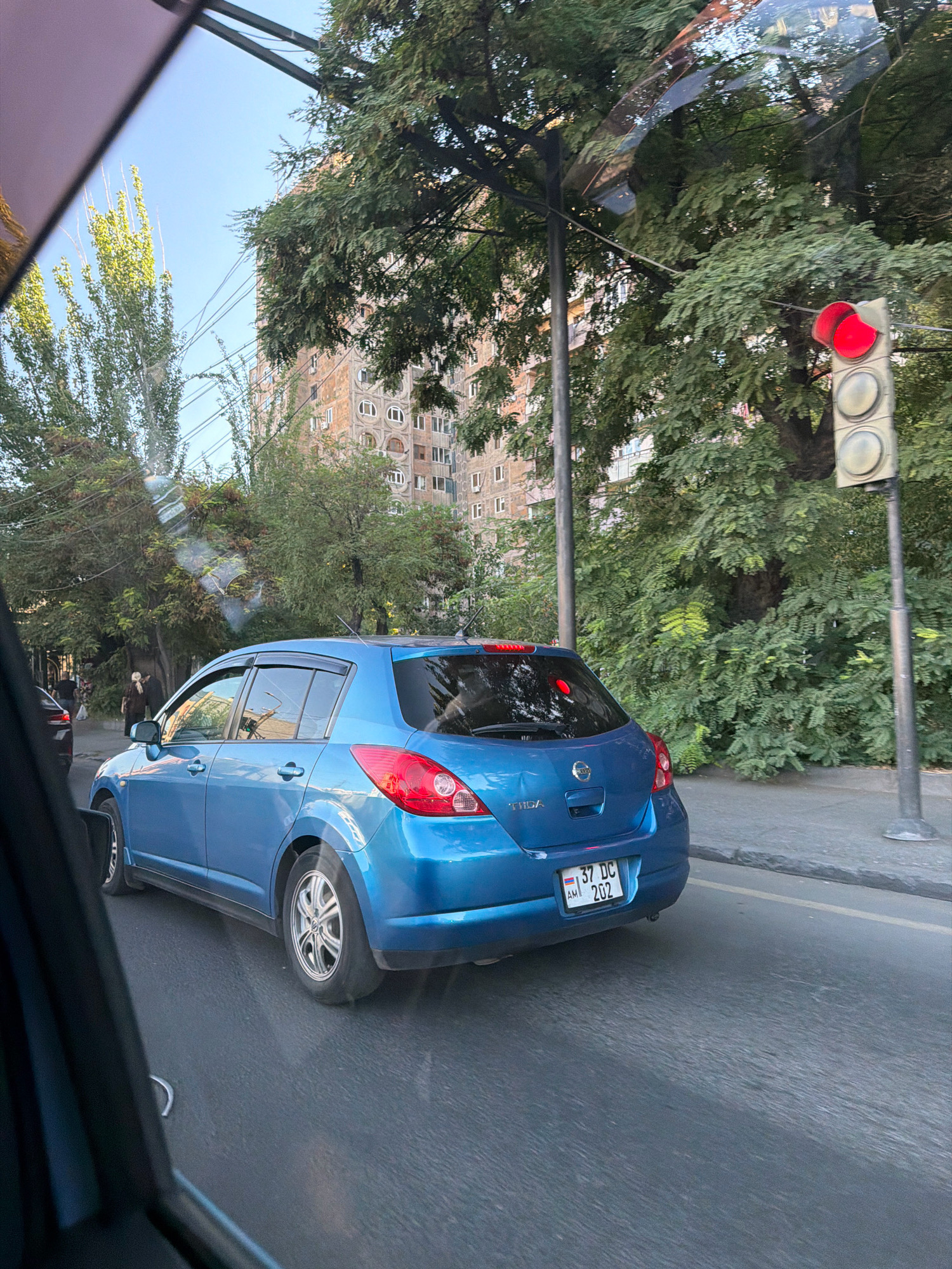 A blue Nissan Tiida is traveling down a street in Yerevan, Armenia. The car has a license plate that reads "37 DC 202 AM". The traffic light is red. Trees line the street, and a tall building with circular windows is visible in the background. The sky is clear and blue. The image appears to be taken from another vehicle.