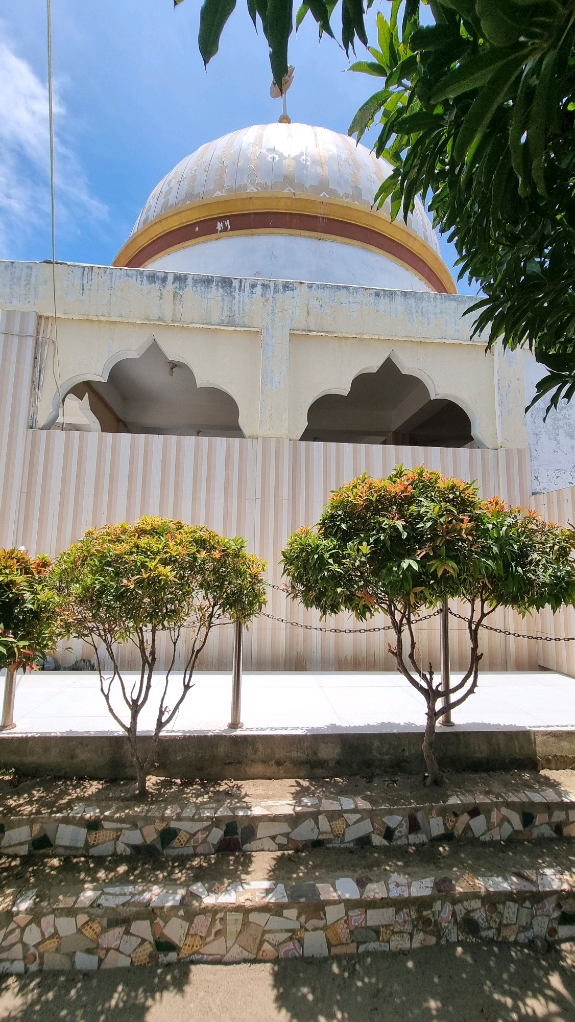 The image shows the exterior of a building with a dome, likely a religious structure, under a bright blue sky with scattered clouds. The building's facade is a light color, possibly cream or white, with decorative arched openings on the upper level. Below these arches, the wall is covered in vertical beige and white stripes. In the foreground, there are two small, neatly trimmed trees with green foliage and reddish new growth. These trees are planted in a paved area, with a low wall and decorative mosaic-covered steps leading down to a shaded area. A thin chain fence separates the paved area from the steps. The dome itself is white with a golden-yellow band and a reddish-brown band just below it. The surface of the dome shows signs of wear and peeling paint. Some green leaves from a larger tree are visible in the upper right corner of the image, casting shadows. The overall impression is of a sunny day in an outdoor area with architectural elements and landscaping. The location is identified as Manetunong, Indonesia.