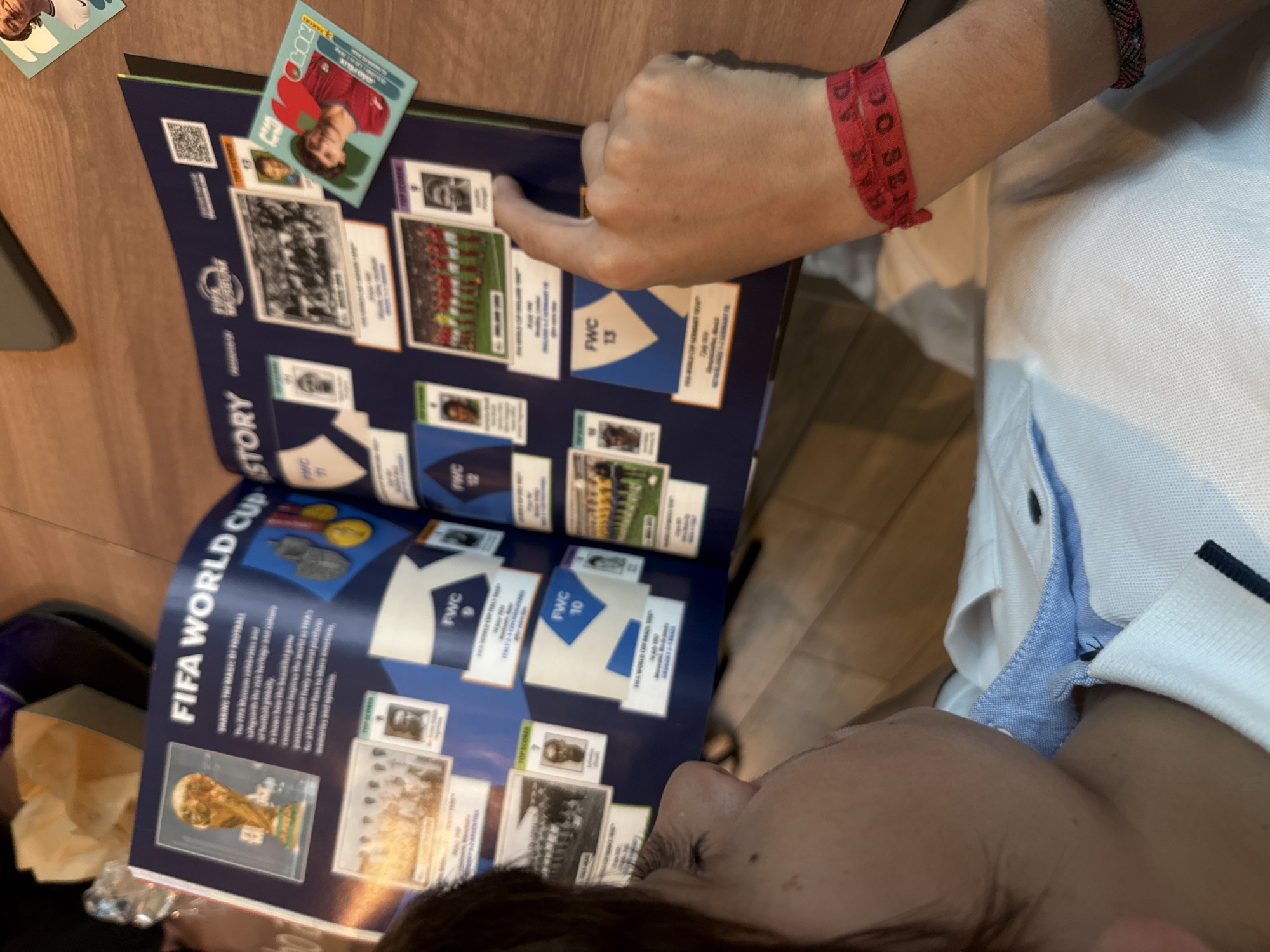 A person is seen indoors, likely in Matosinhos, Portugal, engrossed in a FIFA World Cup sticker album. Their right hand points to a section within the album, while an unpeeled Panini sticker featuring Portuguese footballer João Félix rests on the open page. The individual wears a white polo shirt with blue accents and a red fabric wristband that has faded text, including "DOSE...".
The open sticker album displays pages dedicated to the "FIFA WORLD CUP STORY," featuring sections with text such as "SHARING THE MAGIC OF FOOTBALL" and an explanation of the FIFA Museum's role in preserving football heritage. Various panels are labeled "FWC 6," "FWC 10," "FWC 12," and "FWC 13," corresponding to historical World Cups, including "1966 WORLD CUP ENGLAND," "1970 WORLD CUP BRAZIL," "1974 WORLD CUP GERMANY FR," and "1978 WORLD CUP ARGENTINA." These sections contain small images of teams and players, with some designated "TOP SCORER." A prominent image of the FIFA World Cup trophy and a "FIFA MUSEUM" logo are also visible on the page. A QR code is present in the top left of the album. The activity suggests collecting or reviewing football memorabilia in a casual setting with a wooden surface, possibly a table or floor.