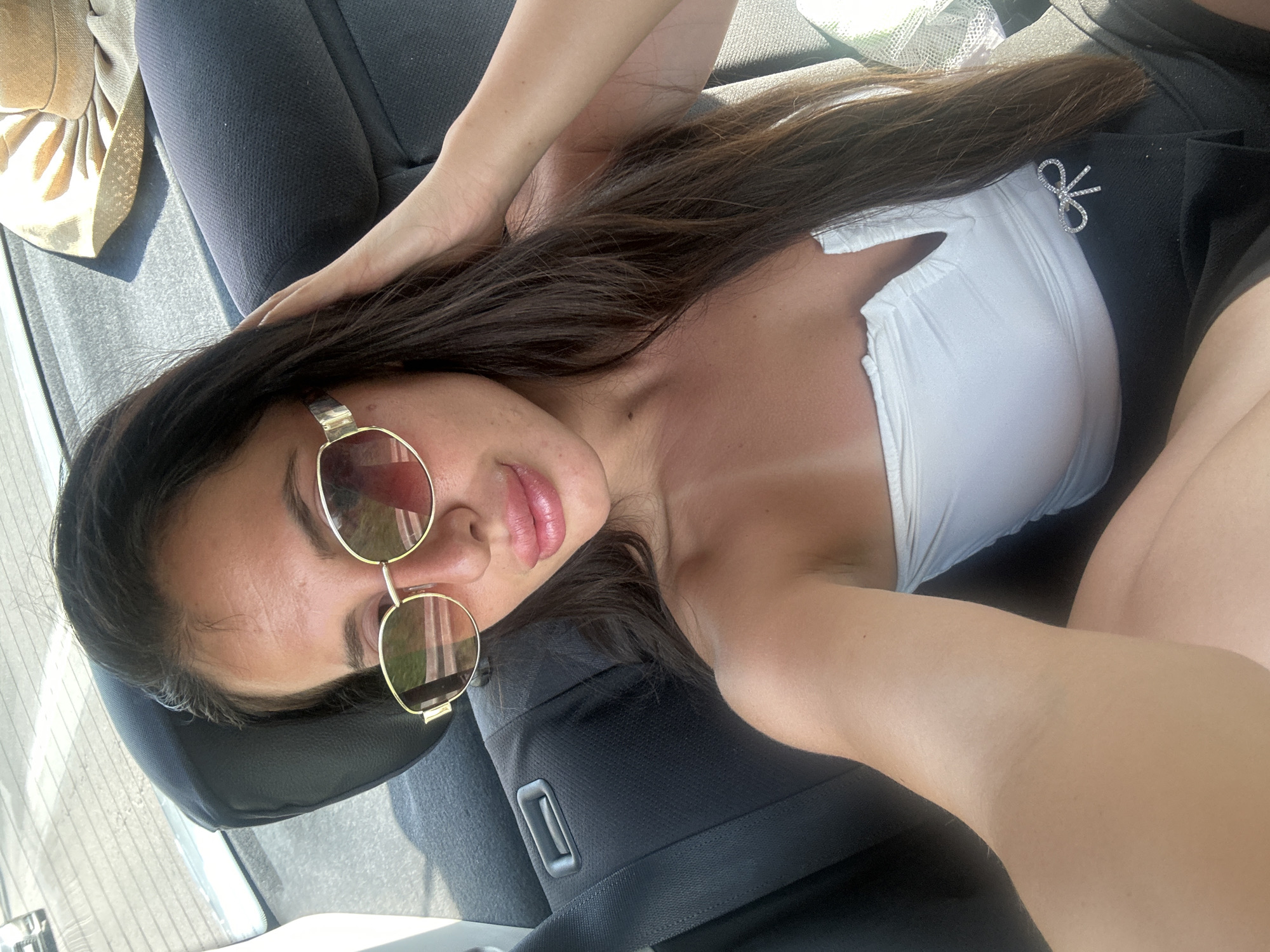 A woman with long, dark hair and sunglasses is taking a selfie in the back seat of a car. She is wearing a white strapless top and a black skirt with a jeweled bow. The car appears to be in motion, as evidenced by the blurred background of a road and other vehicles. There is a straw hat on the seat behind her. The scene is likely taking place during the day, with bright sunlight.
