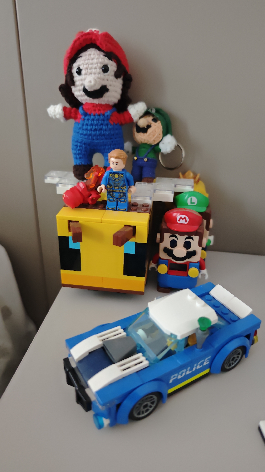 The image shows a collection of toys and figures on a light-colored surface, likely a table or shelf. In the foreground is a blue and white LEGO police car. Behind the car are several figures on top of a yellow block structure, resembling a LEGO Minecraft bee. The figures include: a crocheted Mario and Luigi, a LEGO Star-Lord, a LEGO Mario, and a red fire hydrant.
There is no evidence to suggest this image was taken in Burgas, Bulgaria.