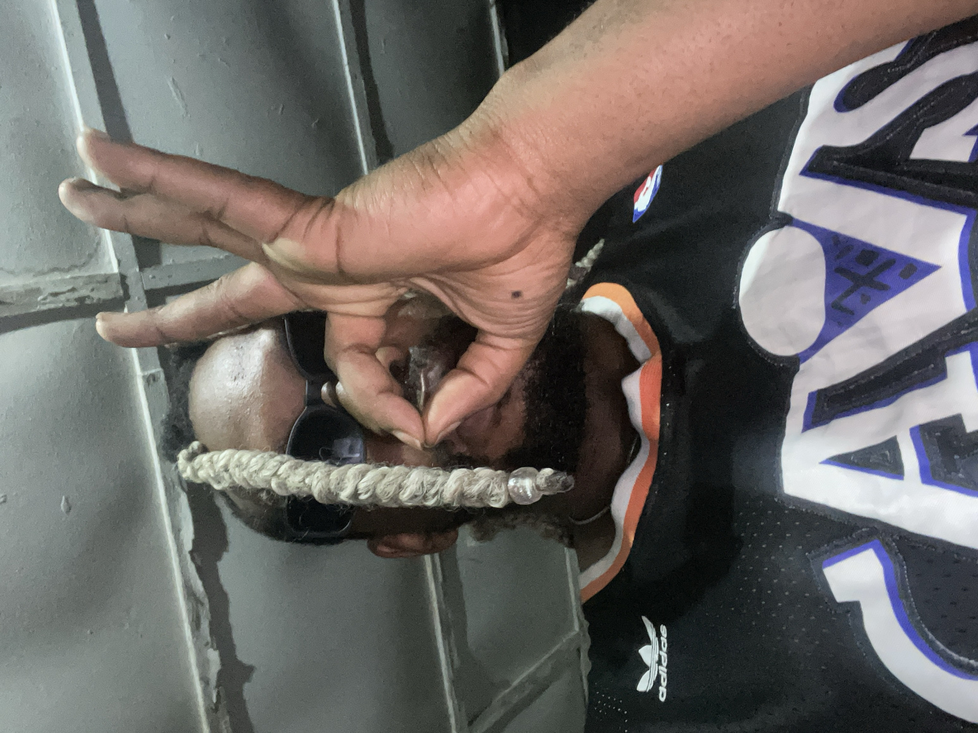 In this close-up, medium shot, a Black man with dreadlocks and sunglasses is shown holding what appears to be a marijuana joint between his index finger and thumb. His right hand is raised to his face, obscuring the joint. The man is wearing a black basketball jersey with orange trim and the word "CAVS" in large white letters outlined in blue. The Adidas logo is visible on the left chest of the jersey. The background consists of a textured, discolored ceiling with exposed conduit. The overall impression is one of casual or clandestine activity, possibly in a dimly lit indoor setting. The image does not provide any specific visual cues to determine its exact location within Jos, Nigeria, beyond the implied urban setting.