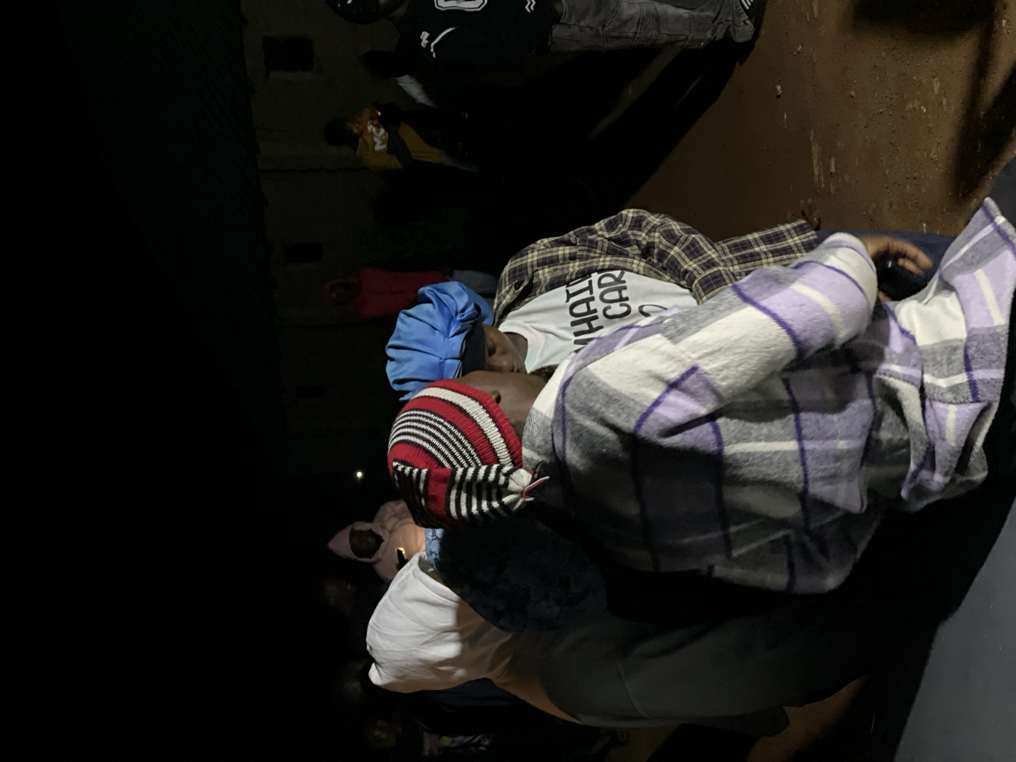In Wumat, Nigeria, at night, a group of young people are gathered outdoors. The ground is dirt. In the foreground, two individuals are facing each other. One person is wearing a gray and purple plaid jacket over a white t-shirt with text that is partially visible. They also have a red and black striped beanie. The second person is wearing a blue head covering and a plaid shirt. Behind them, other individuals are visible, some in hoodies and some holding what appears to be a phone with a light. The background consists of a simple building structure. The lighting is dim, suggesting artificial illumination is present.