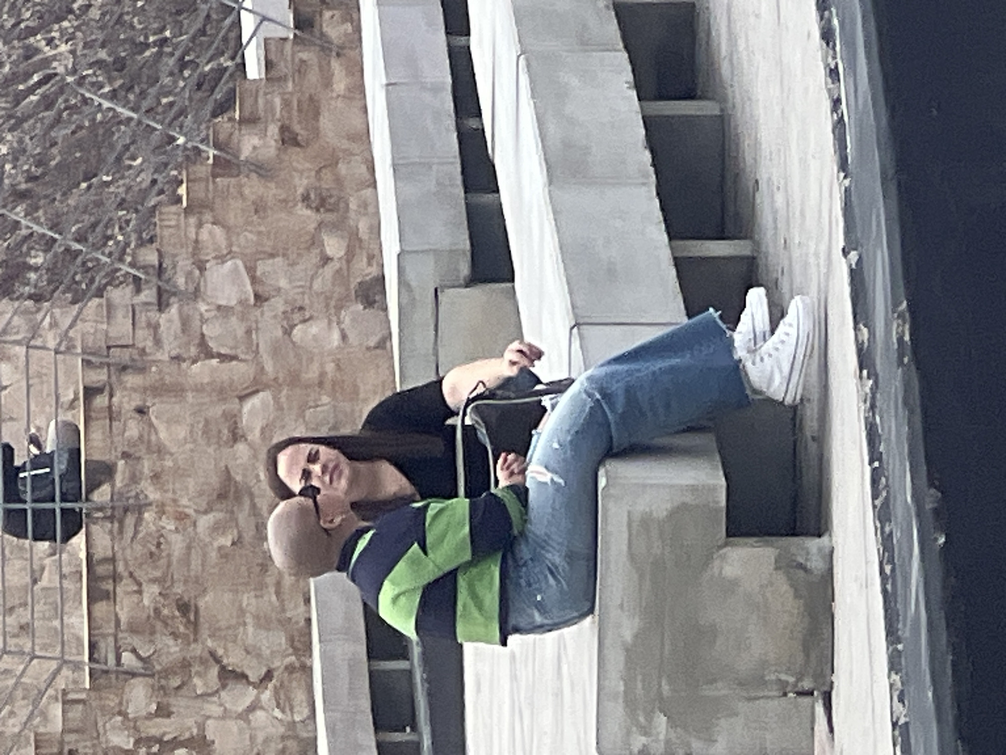 In a daytime scene, two individuals are depicted resting on concrete architectural elements. The image is oriented vertically, but if rotated, it shows one person, bald and wearing a green and navy striped long-sleeve shirt with blue ripped jeans and white sneakers, lying with their legs extended onto a concrete ledge. This person's head is turned towards the second individual. The second person, with long dark hair and wearing a black long-sleeve shirt, is seated upright, holding a dark bag or backpack, and looking at the first person. Their expressions appear calm and engaged, suggesting a casual conversation or interaction.
The background features a textured stone wall, possibly part of an old building or ruin, and on the far left, a metal fence or scaffolding is visible. The immediate surroundings include stark, modern concrete structures, possibly steps or ledges, creating a contrast with the older stonework. The lighting indicates a clear, bright day. There is no visible text in the image. Given the provided context, the setting is in Las Palmas, Spain.