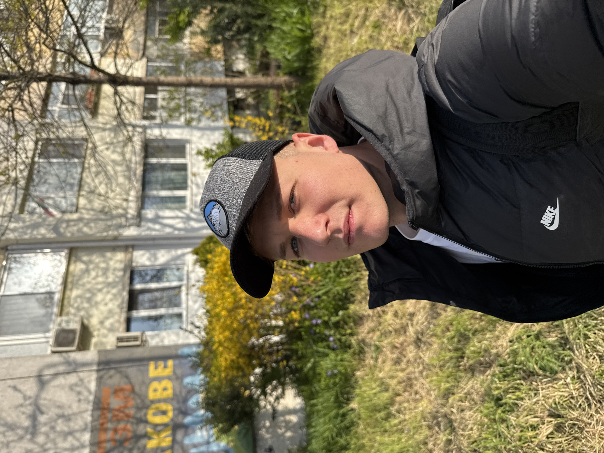 A young man wearing a black Nike jacket and a grey and black cap is facing the camera. He is standing outside on grass. Behind him is a light-colored building. Yellow flowers and green foliage are visible. Part of the word "KOBE" is visible on the lower part of the building, suggesting the location is likely a mural of Kobe Bryant. Based on this information and the architectural style of the building, the location is likely Burgas, Bulgaria, as there is a prominent Kobe Bryant mural in that city.