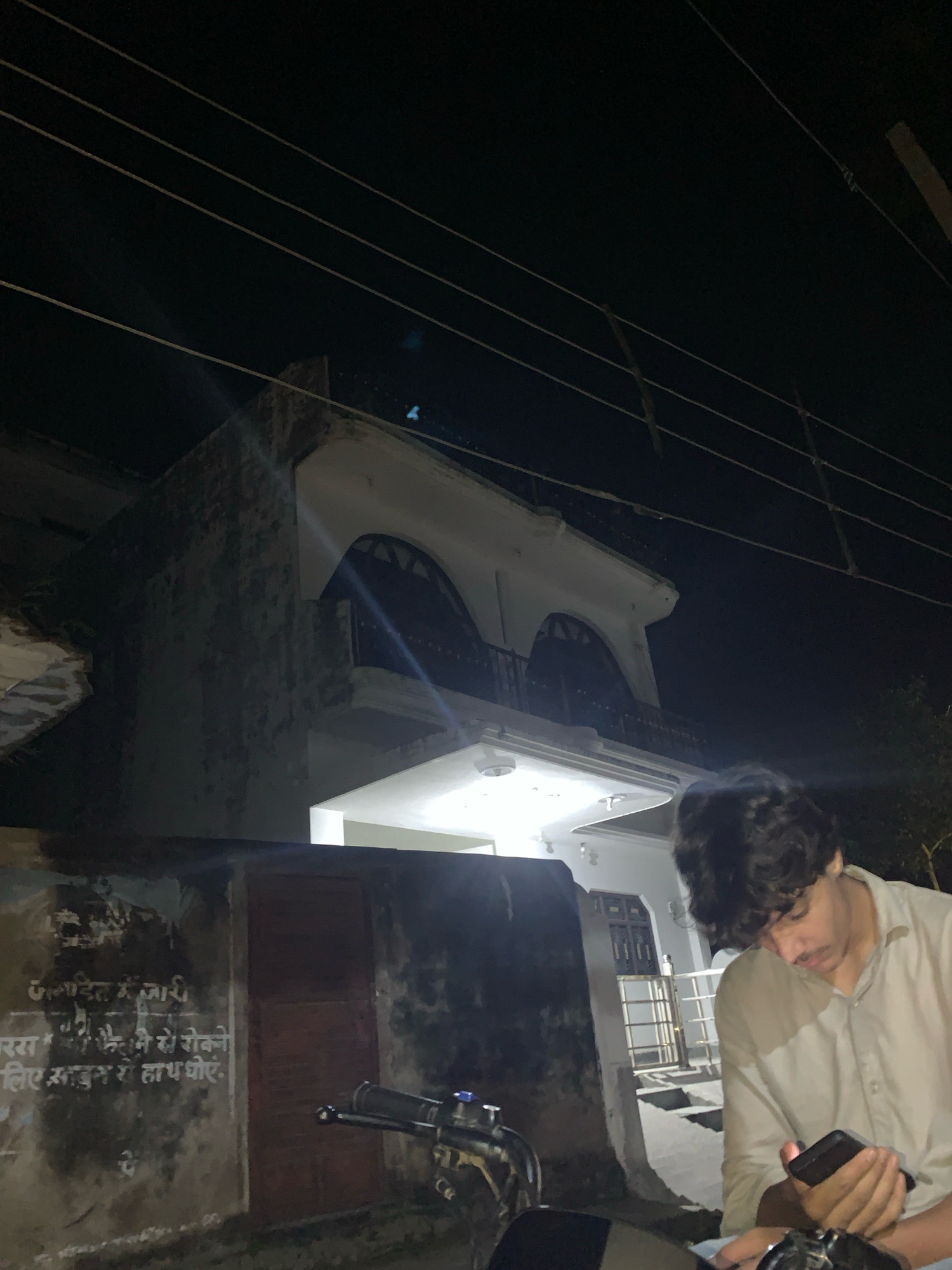 In Parakamal, India, a young man with dark, curly hair is seated on a motorcycle at night, looking down at his mobile phone. He is wearing a light-colored button-down shirt. Behind him, a two-story building with an arched balcony is illuminated by interior lights. The building has a white facade and dark-colored window accents. To the left of the building, a section of a rough, weathered wall is visible, with some faded Hindi text painted on it. The foreground shows the handlebars and front of the motorcycle. Overhead, multiple electrical wires are strung against the dark night sky. The scene appears to be a residential street or courtyard.