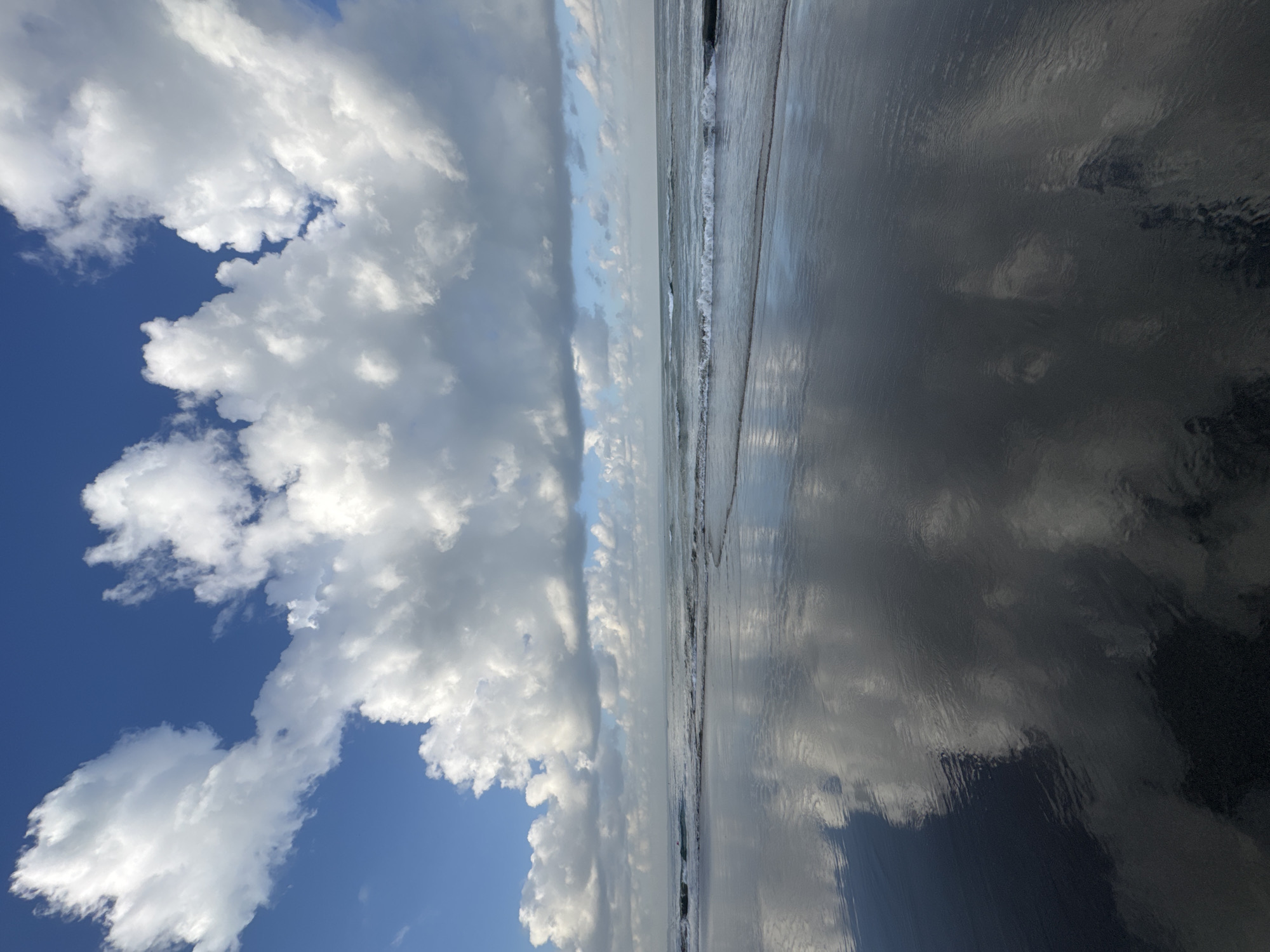 The image depicts a beach scene in North Kuta, Indonesia, under a partially cloudy blue sky. The foreground is dominated by a wet, dark sand beach that reflects the sky and clouds like a mirror. Gentle waves are breaking on the shore, creating a thin line of white foam. In the distance, the ocean stretches to the horizon, meeting the sky. Large, fluffy white clouds are prominent in the sky, with some smaller clouds scattered further back. The overall impression is one of a serene and expansive coastal environment. There are no people or specific objects of interest visible in the scene, just the natural elements of beach, ocean, and sky.