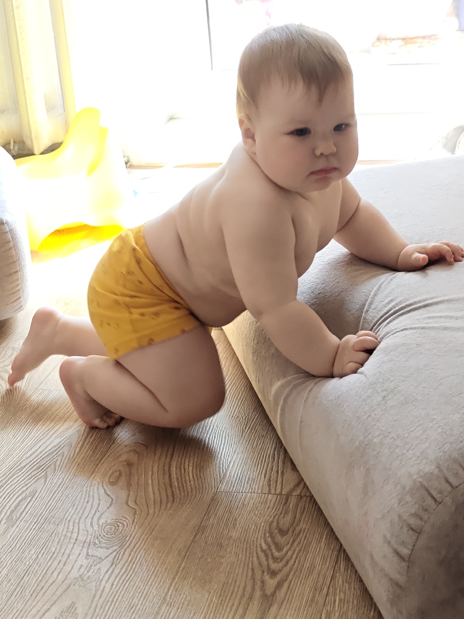 A baby wearing yellow shorts is crawling onto a beige sofa on a wood floor. A yellow object is in the background near a window. The location is the city of Chaikovsky, Russia.