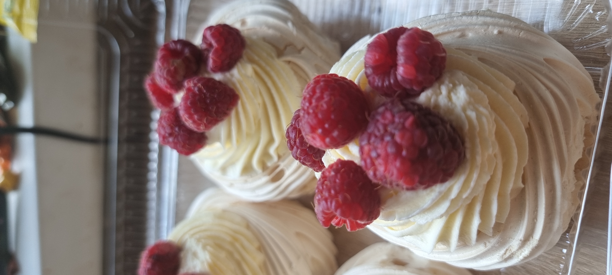 A close-up, eye-level, vertical shot displays three raspberry-topped pavlovas arranged in a clear plastic container. The pavlovas feature swirls of white cream and are garnished with fresh raspberries. The background is blurred, showing hints of a light-colored surface and a dark vertical element. The image appears to be taken indoors, possibly in a bakery or home setting in Aktobe, Kazakhstan, though there are no specific visual cues to confirm this location. The lighting is soft and natural.