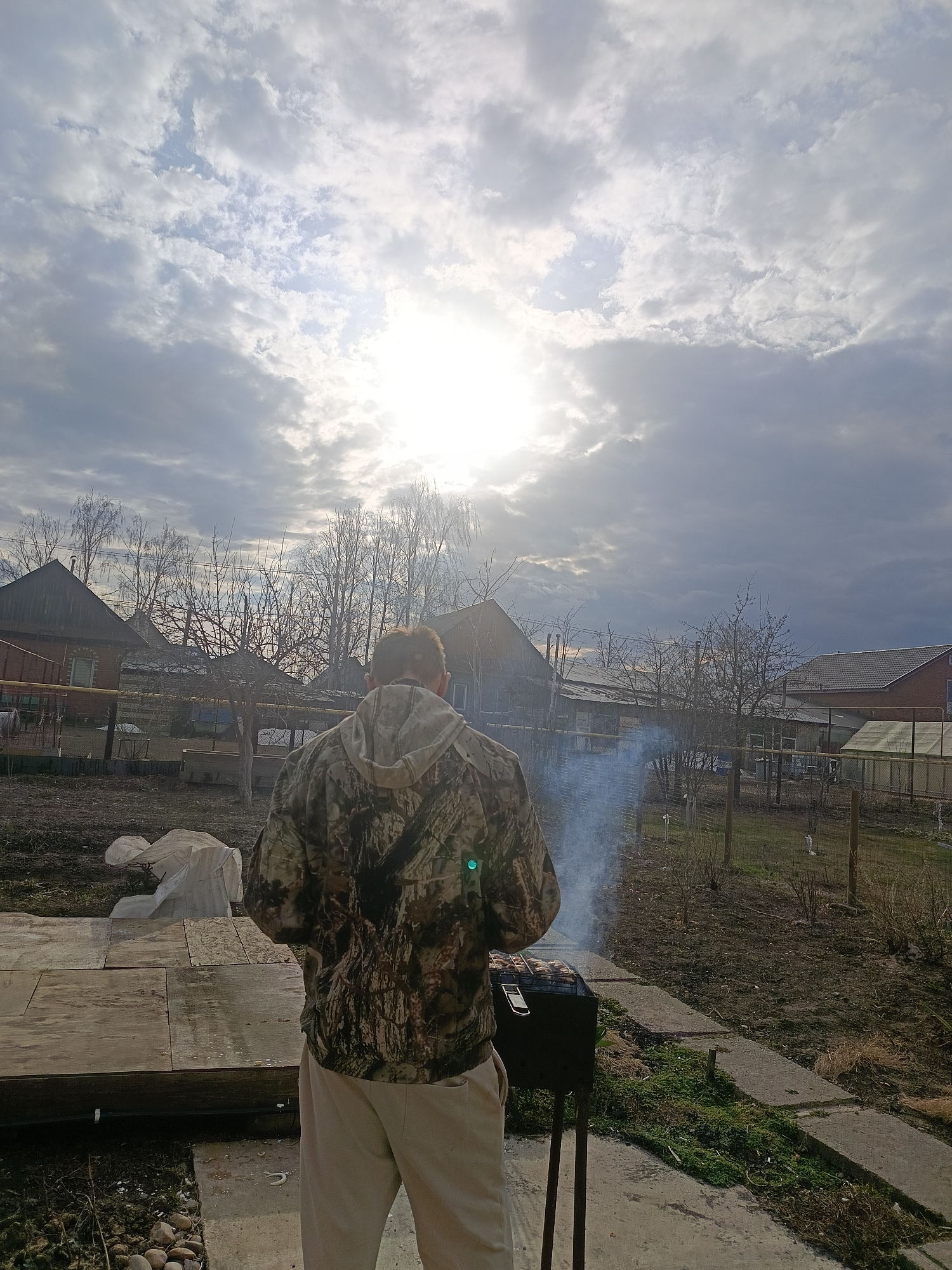 An outdoor scene in Chaikovsky, Russia, captures a person grilling under a cloudy yet brightly lit sky. The individual, seen from the back, is wearing a camouflage-patterned hooded jacket and light-colored trousers. They are standing on a concrete surface, facing a black, portable grill from which a plume of white smoke ascends, indicating cooking activity. Some food is visible on the grill grate.
The setting is a rural or suburban backyard, possibly a dacha plot. The background features several houses, varying in construction from brick to wood, with dark-tiled or sheet roofs. Numerous bare deciduous trees are scattered throughout the landscape, suggesting the season is late winter or early spring. The ground is uneven, with patches of dry grass, moss, and exposed earth, alongside concrete paths and a wooden platform in the foreground to the left. A white tarp or covering lies on the ground near the wooden structure. Fences are also visible, delineating property lines.
The sky is largely covered with a mix of grey and white clouds, but the sun shines intensely from the upper right, creating a strong glare and lens flare that illuminates the scene despite the cloud cover. The overall impression is of a casual outdoor cooking event on a day with changeable weather. No visible text is present in the image.