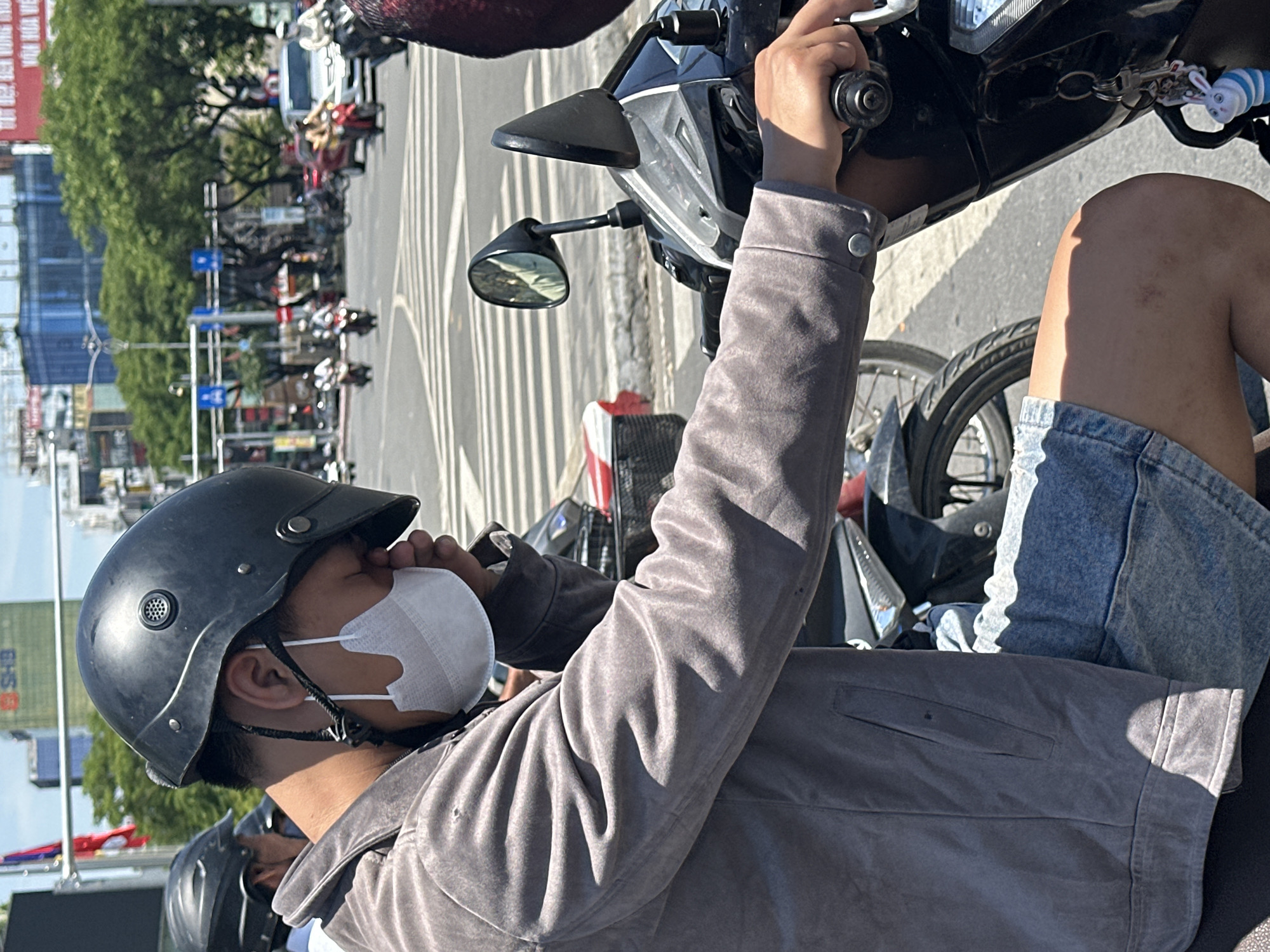 A person wearing a black helmet, a white mask, and a grey jacket is sitting on a motorcycle at a crosswalk in Đà Nẵng, Vietnam. The person's eyes are closed, and they appear to be resting or uncomfortable. The background shows a busy city street with other motorcycles and cars, buildings, and trees. The sun is shining brightly, casting shadows on the road. The image captures a moment of pause in the midst of urban activity, highlighting the individual's personal space amidst the bustling city environment.
