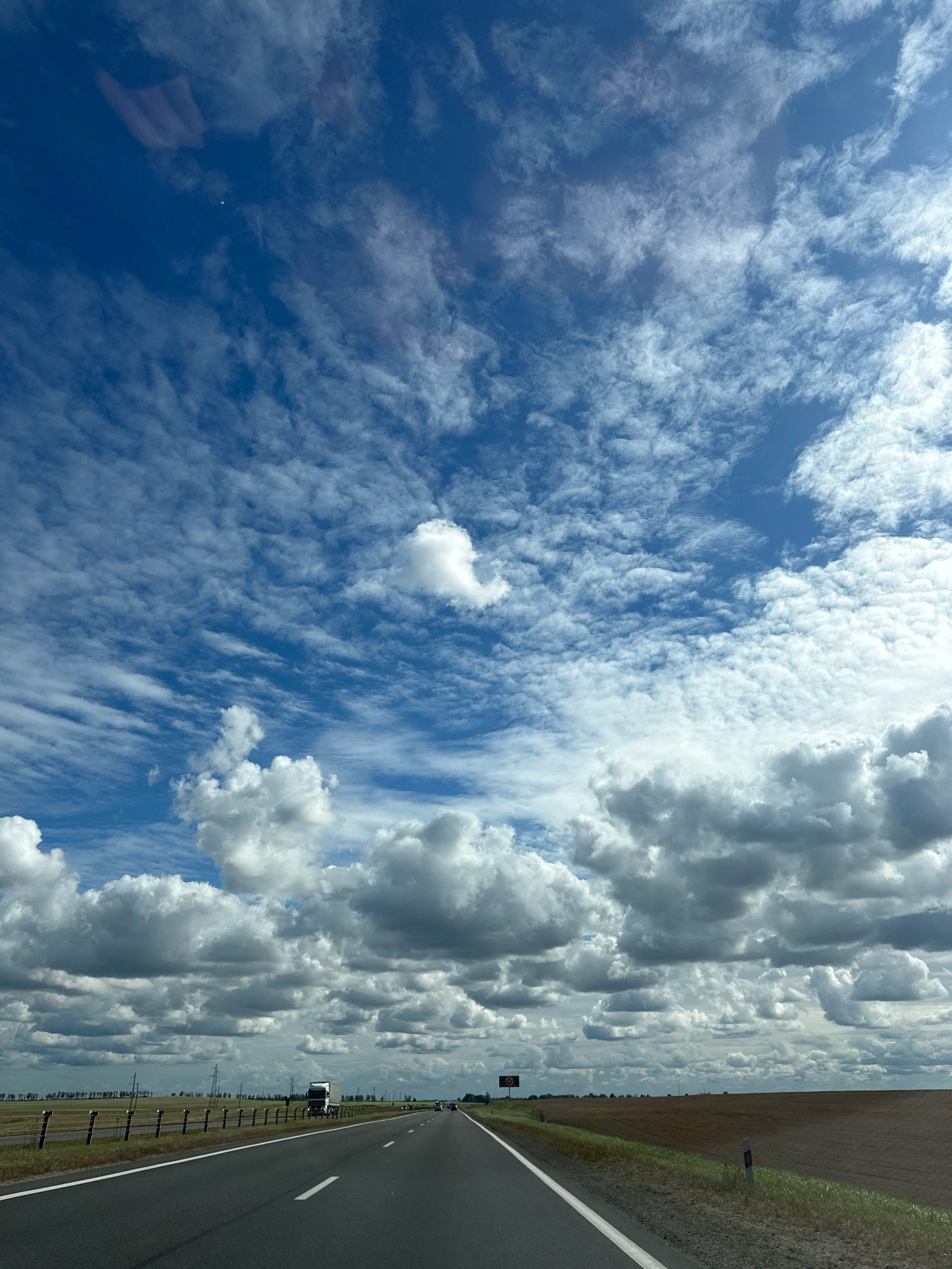 The image shows a view from a vehicle driving on a highway in Турынскі сельскі Савет, Belarus. The highway is asphalt with white lane markings and stretches into the distance. On the left side of the road, there is a fence and then a field with power lines and trees in the background. On the right side of the road, there is a field of harvested crops. The sky is filled with a mix of white cumulus clouds and wispy cirrus clouds against a bright blue sky. A large truck is visible on the highway in the distance, heading in the same direction as the viewer.