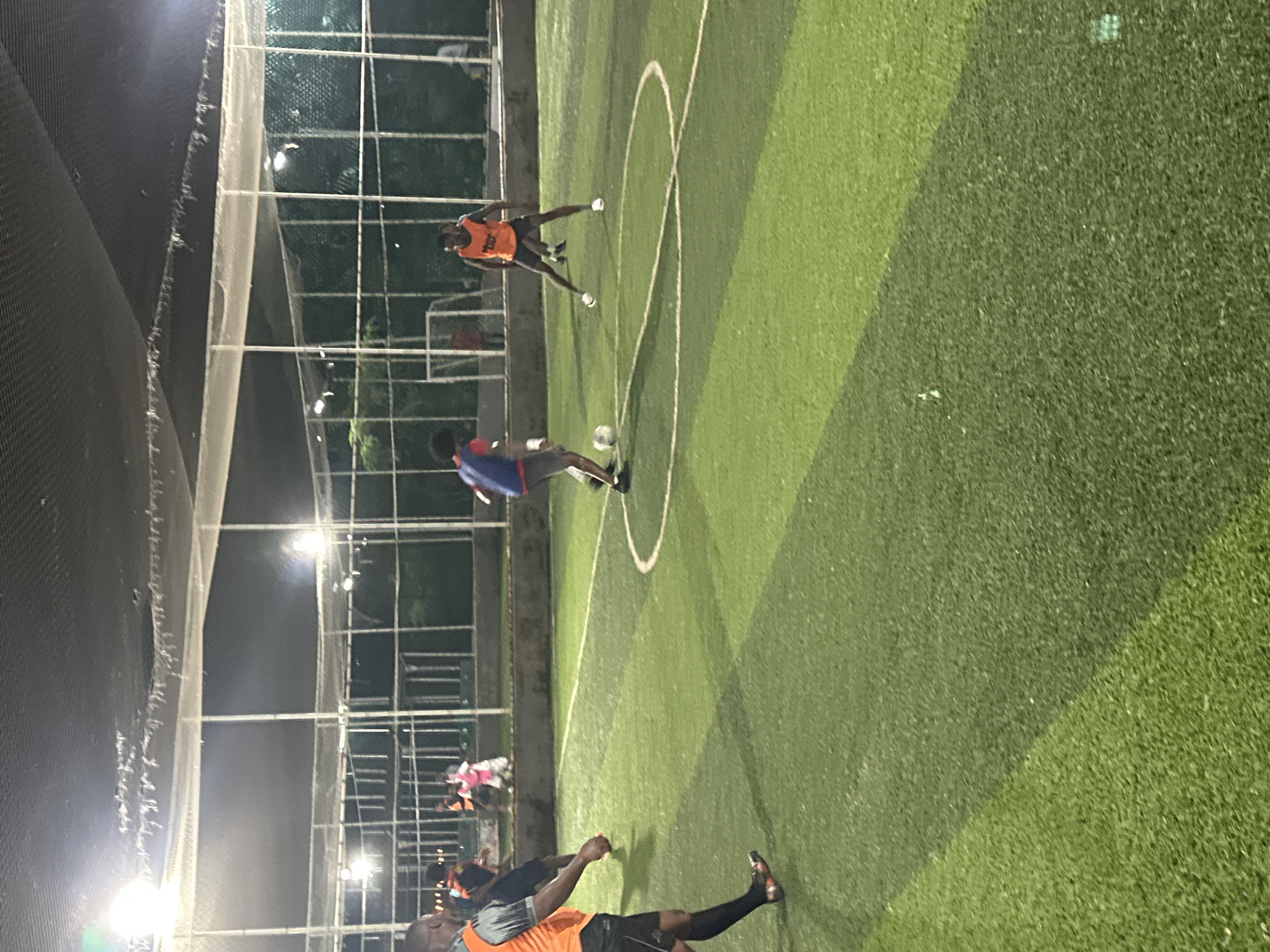 In Abuja, Nigeria, on a brightly lit artificial turf football pitch enclosed by netting and metal poles, a group of individuals are engaged in a game of football. Three players are prominently visible in the foreground and mid-ground. To the left, a man wearing an orange vest with grey shoulders and black shorts is partially visible from the side, facing away from the camera, with his right leg extended. In the center, a player in a blue and red jersey, with black shorts, is actively dribbling a white football with his left foot. To the right, another player in an orange vest and black shorts is positioned in a defensive stance, with his legs spread wide, looking towards the ball. In the background, several other figures are scattered across the pitch, some appearing to be spectators or participants on the sidelines. The lighting suggests the game is taking place in the evening or at night.