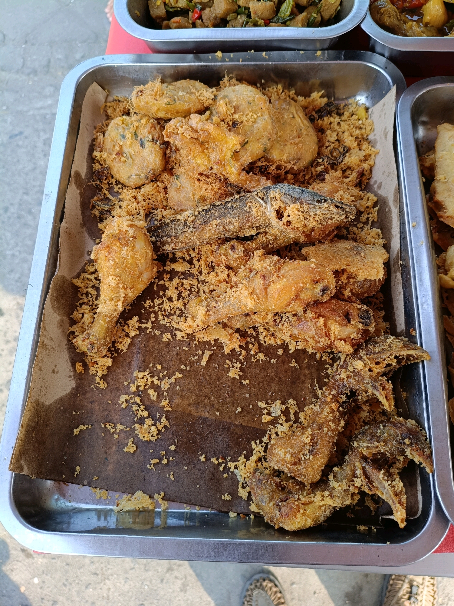 In Maguwoharjo, Indonesia, a metal tray is filled with a variety of fried foods, including chicken pieces, fish, and fritters, all coated in a crispy batter. The food is arranged on brown paper lining the tray. In the background, other trays of food are visible, suggesting a food stall or market setting. The ground beneath the tray appears to be paved.