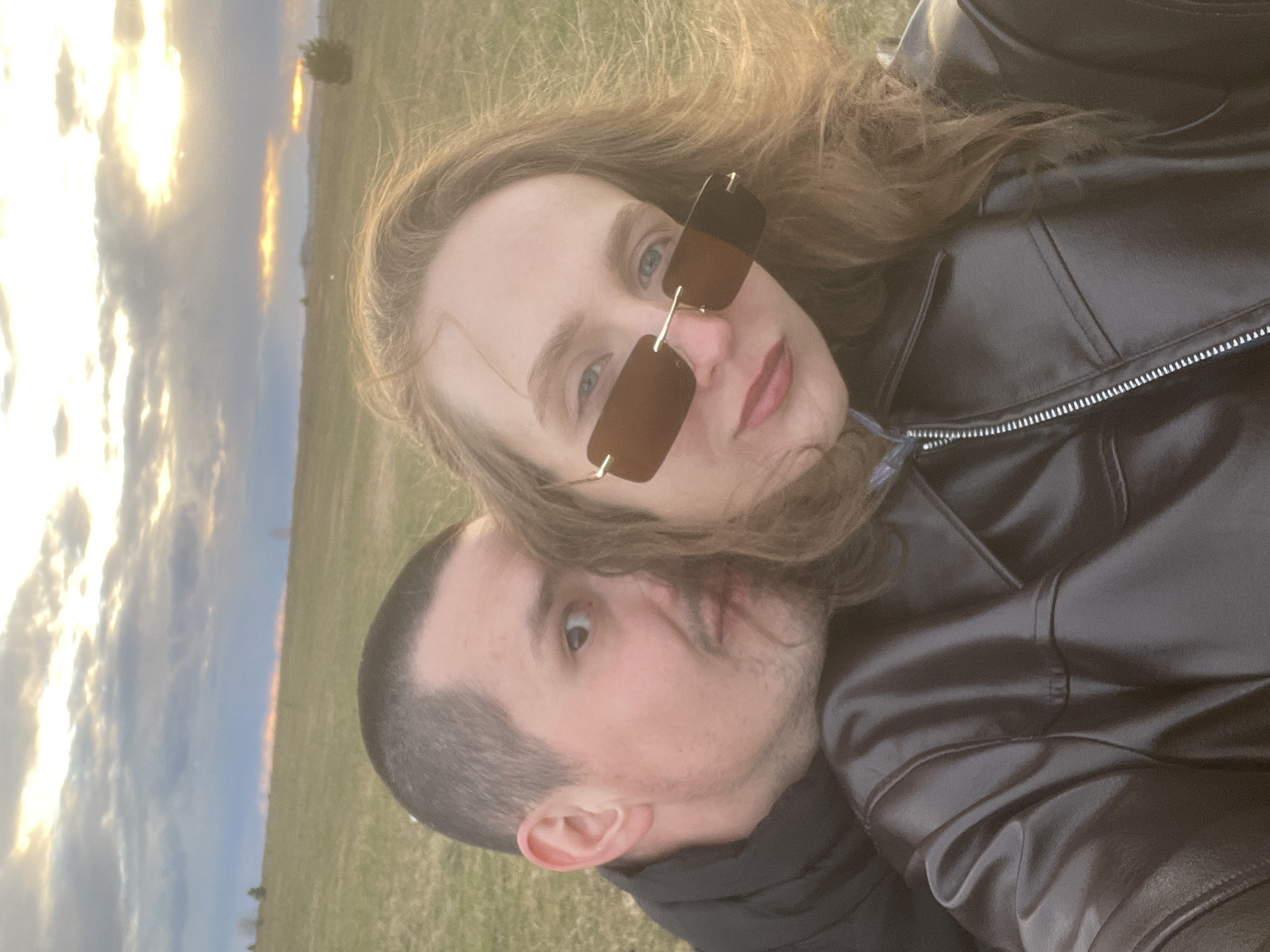 The image features two individuals, a woman and a man, in a close-up outdoor portrait. The photo appears to have been taken during golden hour (sunrise or sunset) in Kueda, Russia, set against a backdrop of a grassy field and a dramatically lit sky.
The woman is positioned in the foreground, slightly above and in front of the man. She has long, wavy light brown hair and is wearing rectangular brown-tinted sunglasses with a delicate gold-colored frame, partially obscuring her blue eyes. Her lips are painted a reddish-pink, and she looks directly towards the viewer with a neutral to slightly smiling expression. She is dressed in a black jacket, possibly leather, which has a visible silver zipper.
The man is situated behind and slightly below the woman. He has short, dark hair and stubble, with his ear prominent. He looks upwards and to his right (from his perspective) with an expression that appears somewhat surprised or curious. He is also wearing a black jacket.
The background shows an expansive sky with scattered clouds, intensely illuminated by the low sun. This creates warm golden and orange hues, particularly along the horizon line. A faint, distant tree can be seen against the brightly lit sky. The setting appears to be an open, grassy field. The overall impression is a casual and intimate shared moment outdoors. No text is visible in the image.
