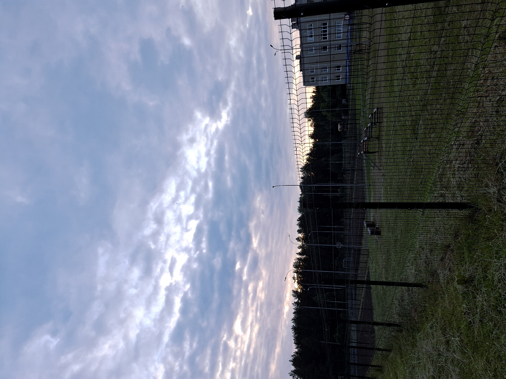 The image depicts an outdoor scene, likely at dawn or dusk, given the lighting. In the foreground, there is a wire mesh fence that runs diagonally across the frame from the bottom left to the top right. Beyond the fence, a grassy area is visible, with a few benches and some playground equipment. In the middle ground, a dense line of dark trees forms a backdrop, and beyond the trees, a light-colored building with multiple windows is visible. The sky above is filled with a mix of clouds, with some areas of light filtering through, suggesting the sun is low on the horizon. The overall impression is of an urban or suburban perimeter area. The location is identified as Марковский, Russia. No people are present in the image, and no specific actions or interactions are depicted.