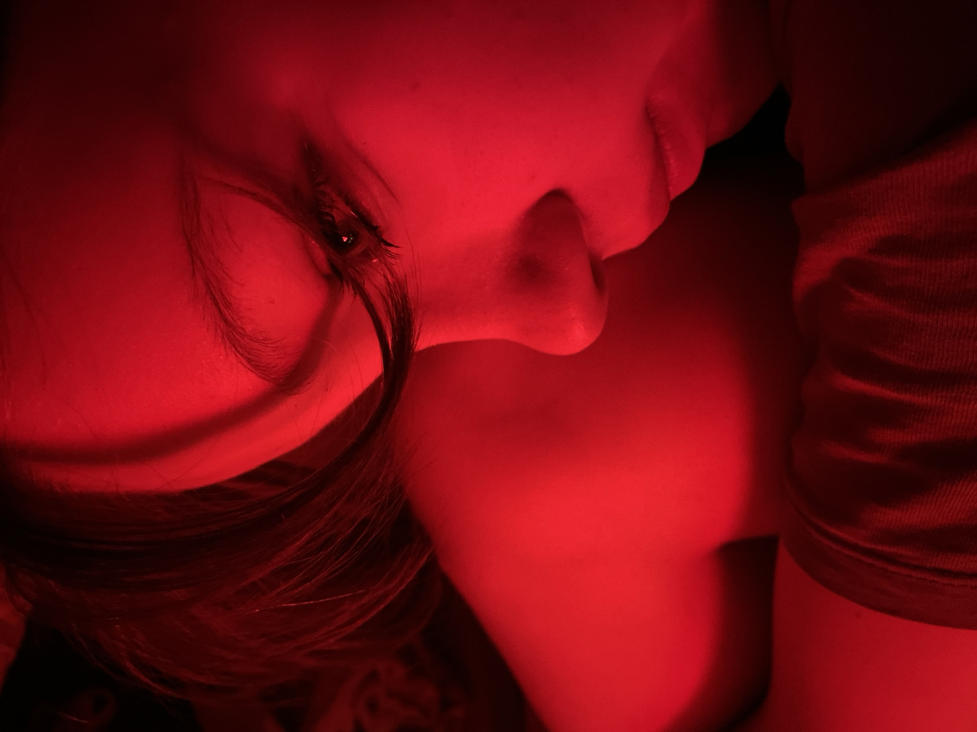 A close-up, low-angle portrait photograph shows a person with fair skin illuminated by a strong red light. The individual is oriented with their face partially visible, the side of their nose, mouth, and one eye visible. Their dark hair is styled and partially obscures their forehead. The background is obscured by the intense red light. The subject also appears to be wearing a red shirt. The photograph does not contain information to determine if the image was taken in the city of Chaikovsky, Russia.