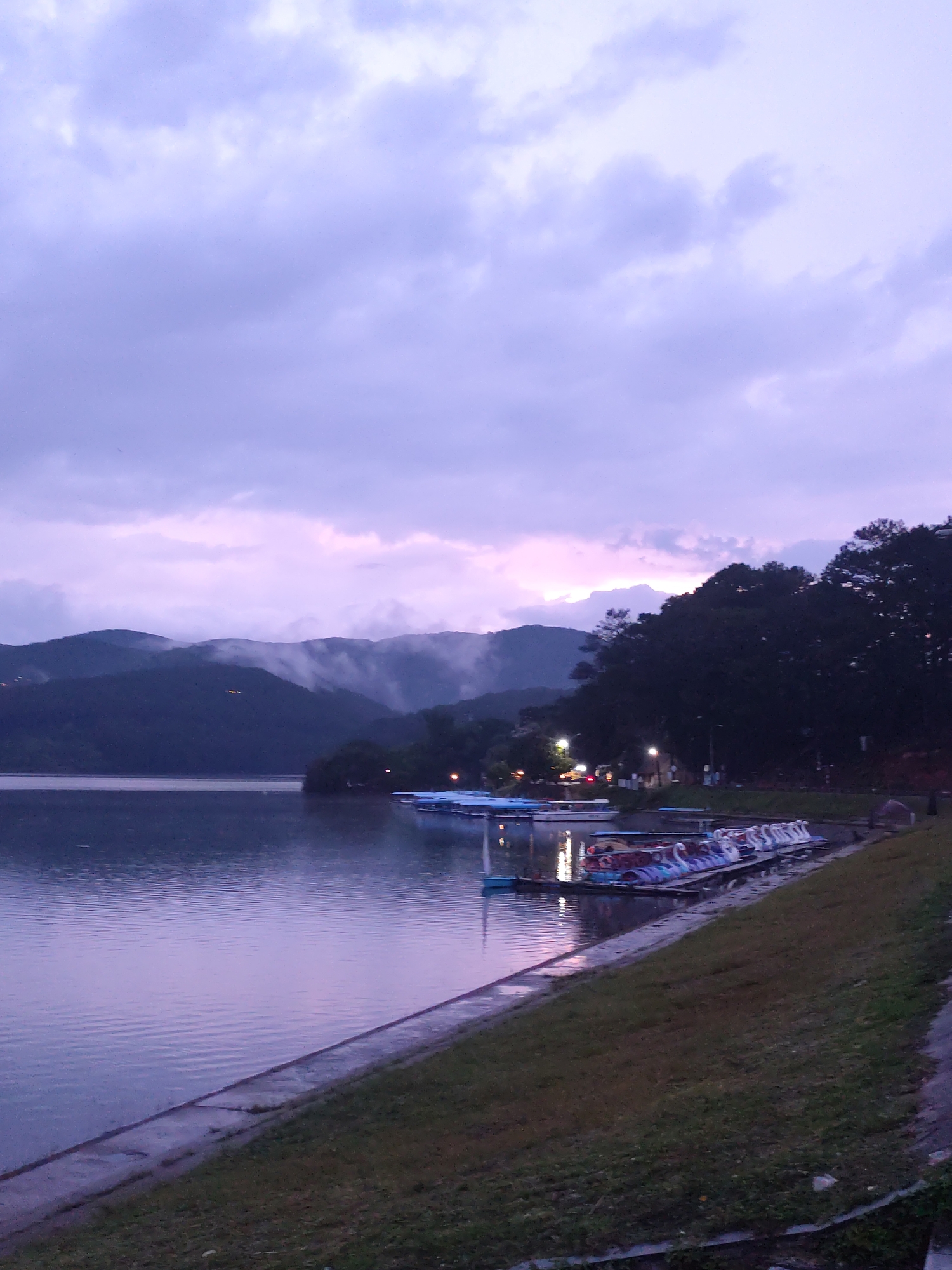 The image depicts a lakeside scene at dusk or dawn in Da Lat, Vietnam. The sky is filled with purplish clouds, and mountains are visible in the background, some shrouded in mist. The water is calm and reflects the sky's colors. A concrete embankment separates the water from a grassy area on the right. A dock extends into the water, holding several swan-shaped paddle boats. Lights are visible along the shoreline in the distance, suggesting a populated area. There are no people clearly visible in the immediate foreground.