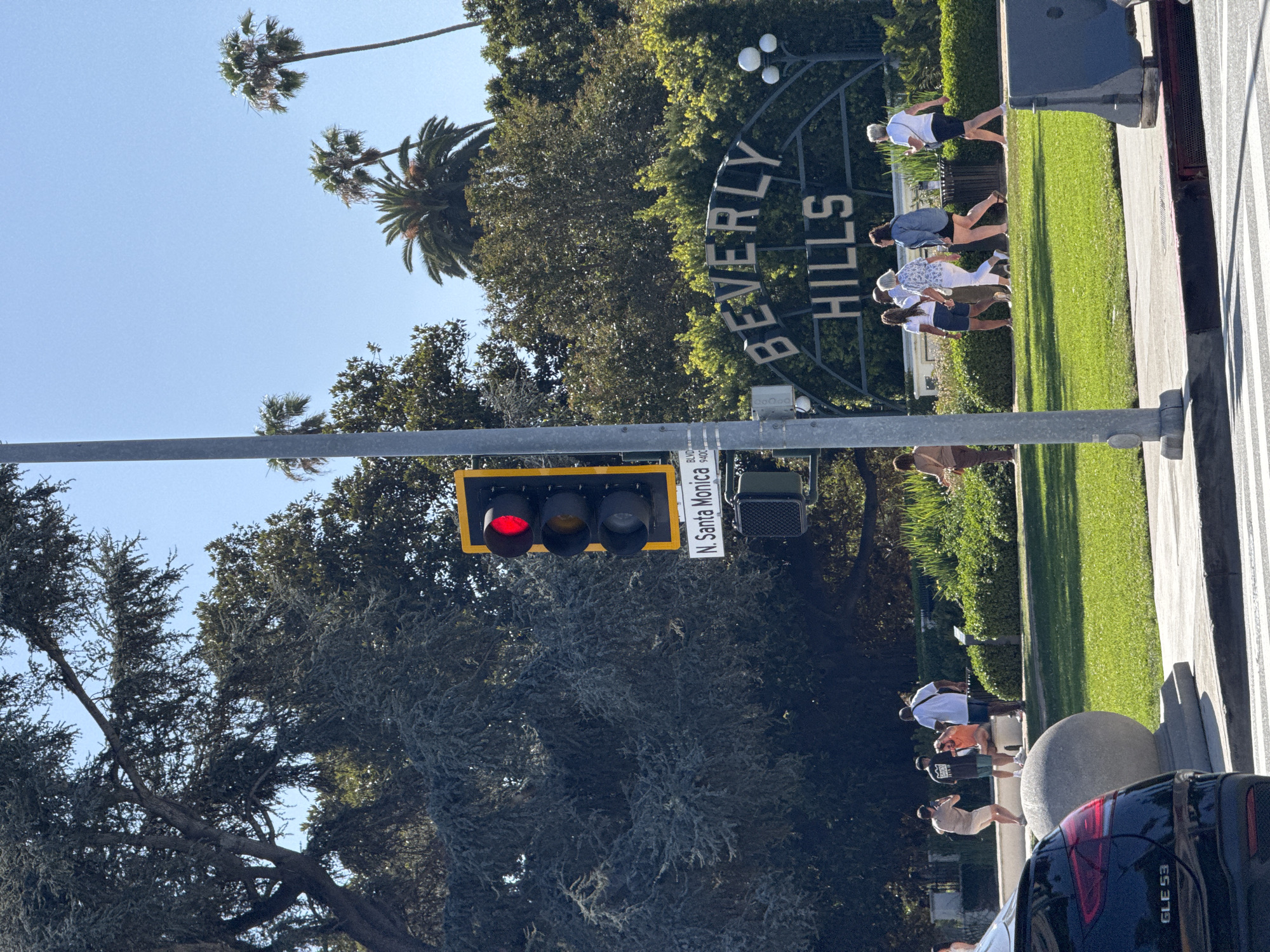 The image shows a daytime view in Beverly Hills, United States of America. A traffic light displays a red signal. A street sign indicates the location as N. Santa Monica Blvd 9400C. In the background, the iconic "Beverly Hills" sign is visible with a group of people standing near it. Some people are taking photos of the sign. Trees and grass surround the area. A black SUV is parked in the foreground.