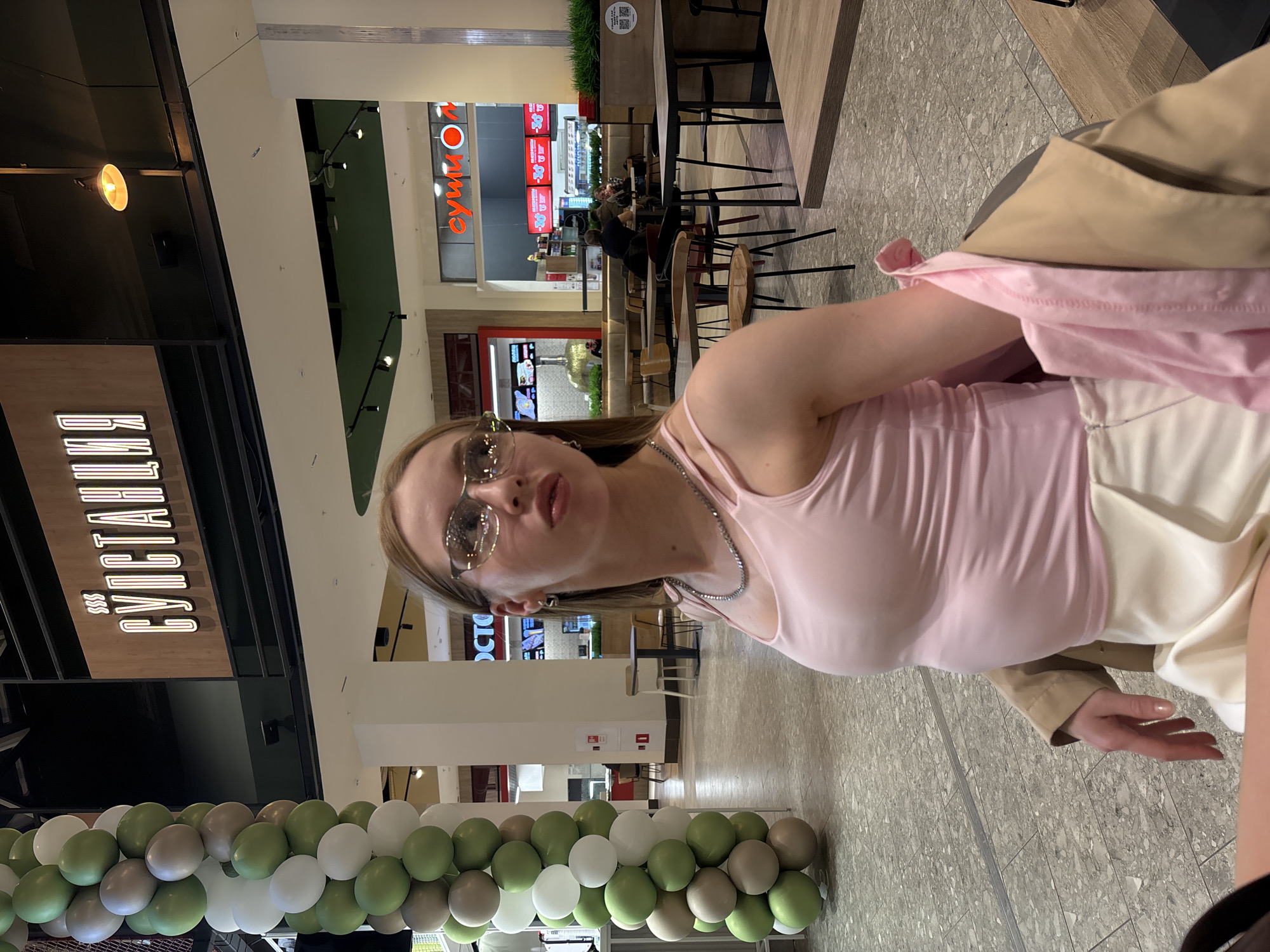 The image captures a young woman posing for a photo in an indoor setting, likely a shopping mall or a large food court in Perm, Russia. The woman is positioned on the right, looking directly at the camera with a slightly pouty expression and puckered lips. She has light brown hair, is wearing reflective sunglasses, a light pink sleeveless top, and a silver-colored chain necklace. A beige or light brown jacket is draped over her shoulders, and her right hand is visible near her hip.
The background shows a spacious, well-lit commercial interior with a tiled floor. To the left, a large illuminated sign clearly reads "БУЛЬВАР" (Boulevard), suggesting a cafe or restaurant. Further back and to the right, another sign with red text on a white background reads "Суши Оn," indicating a sushi establishment. Several people are visible in the mid-ground, seated at tables and appearing to be dining. The furniture includes modern tables and chairs with wooden and metal components. A long decorative arrangement of green, white, and grey balloons extends across the lower-left portion of the frame. A QR code sticker is visible on a planter box in the background. The scene suggests a busy daytime or evening environment within a modern commercial complex.