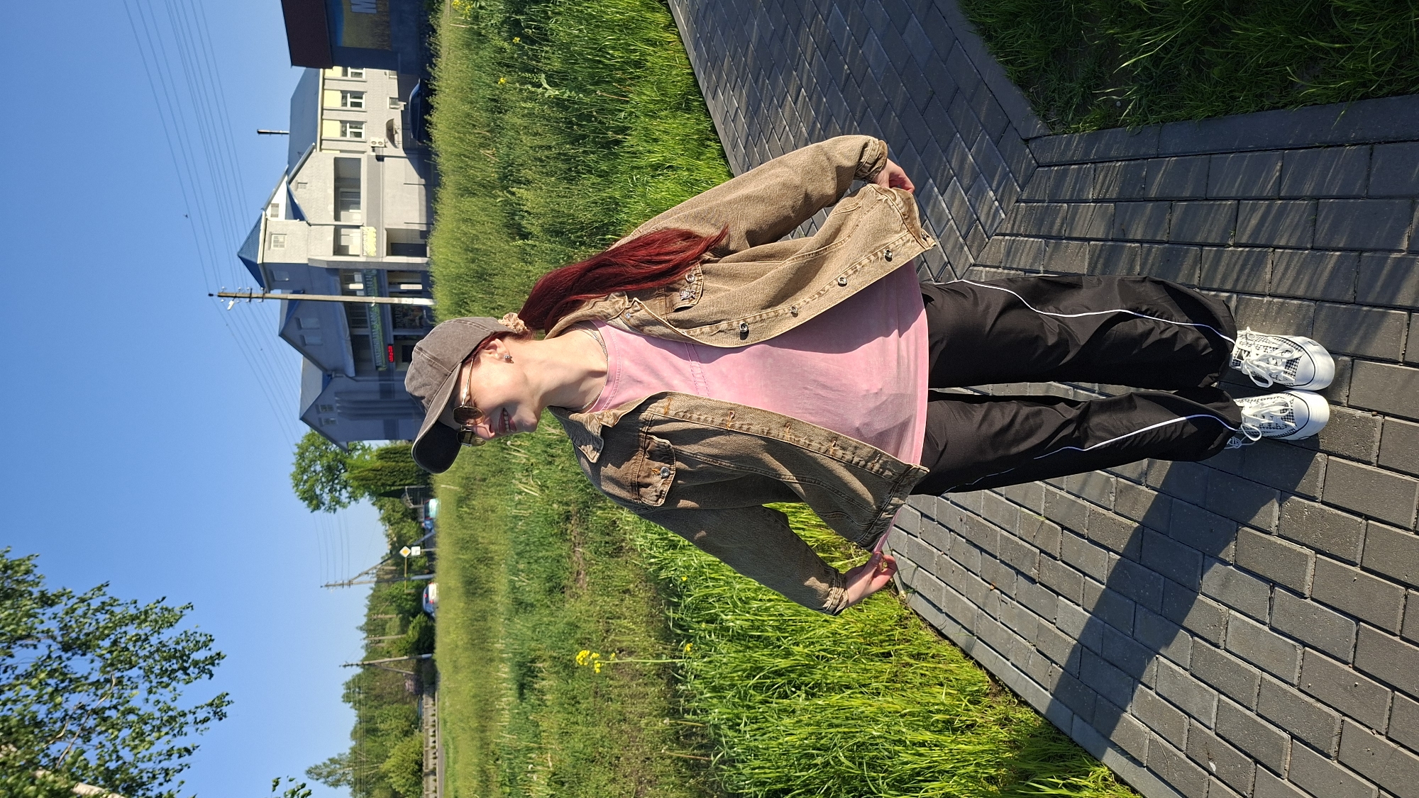 A medium shot depicts a person, likely female, standing on a brick path, likely in Забалоцкі сельскі Савет, Belarus, given the context. She's dressed in black pants with a white stripe down the side, white sneakers, a pink t-shirt, and a brown denim jacket. She's wearing a baseball cap and sunglasses. The background includes green grass, some buildings, and a clear blue sky. The photo is taken during the daytime.