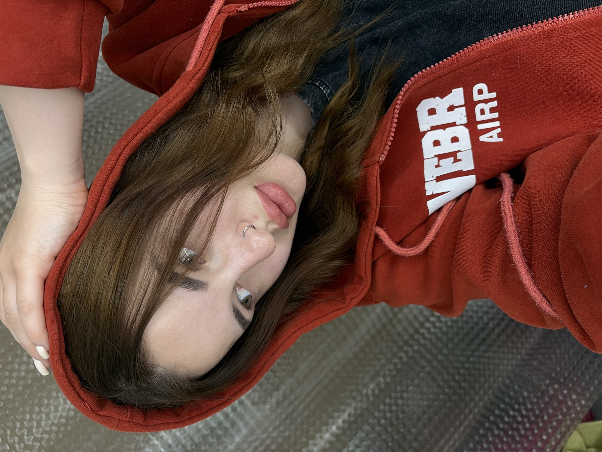 A woman with long brown hair and fair skin is wearing a red hoodie. She is looking down and slightly to the left. The hoodie has white lettering on the right side of the chest. She is wearing a nose ring on her left nostril. The background is a metallic, textured surface. The location is Krasnodar, Russia.