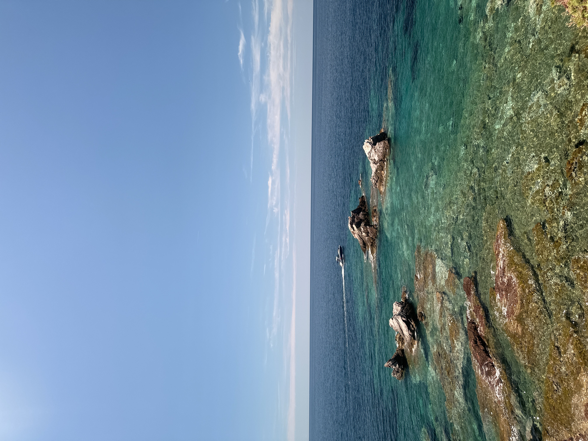 The image depicts a bright, sunny day at the coast near Sveti Stefan, Montenegro. The foreground features shallow, clear turquoise water revealing submerged rocks and a rocky shoreline. Further out, darker blue water stretches to the horizon under a clear blue sky with a few wispy white clouds. A white motorboat is visible in the mid-ground, creating a wake as it moves across the water. Several large rocks break the surface of the sea, some clustered together, others more isolated. The overall scene is one of a serene coastal landscape on a warm, clear day.