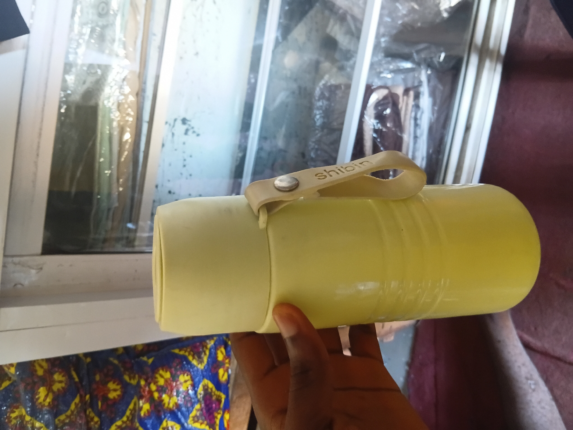 A person's dark-skinned hand is holding a light yellow thermos bottle in front of a glass display case. The thermos has a light beige strap attached to its side with a silver rivet. The word "shibin" is embossed on the strap. The display case contains various items wrapped in plastic, and a colorful patterned fabric is visible to the left of the thermos. The scene appears to be an indoor setting, possibly a shop or market stall, in Jalingo, Nigeria.