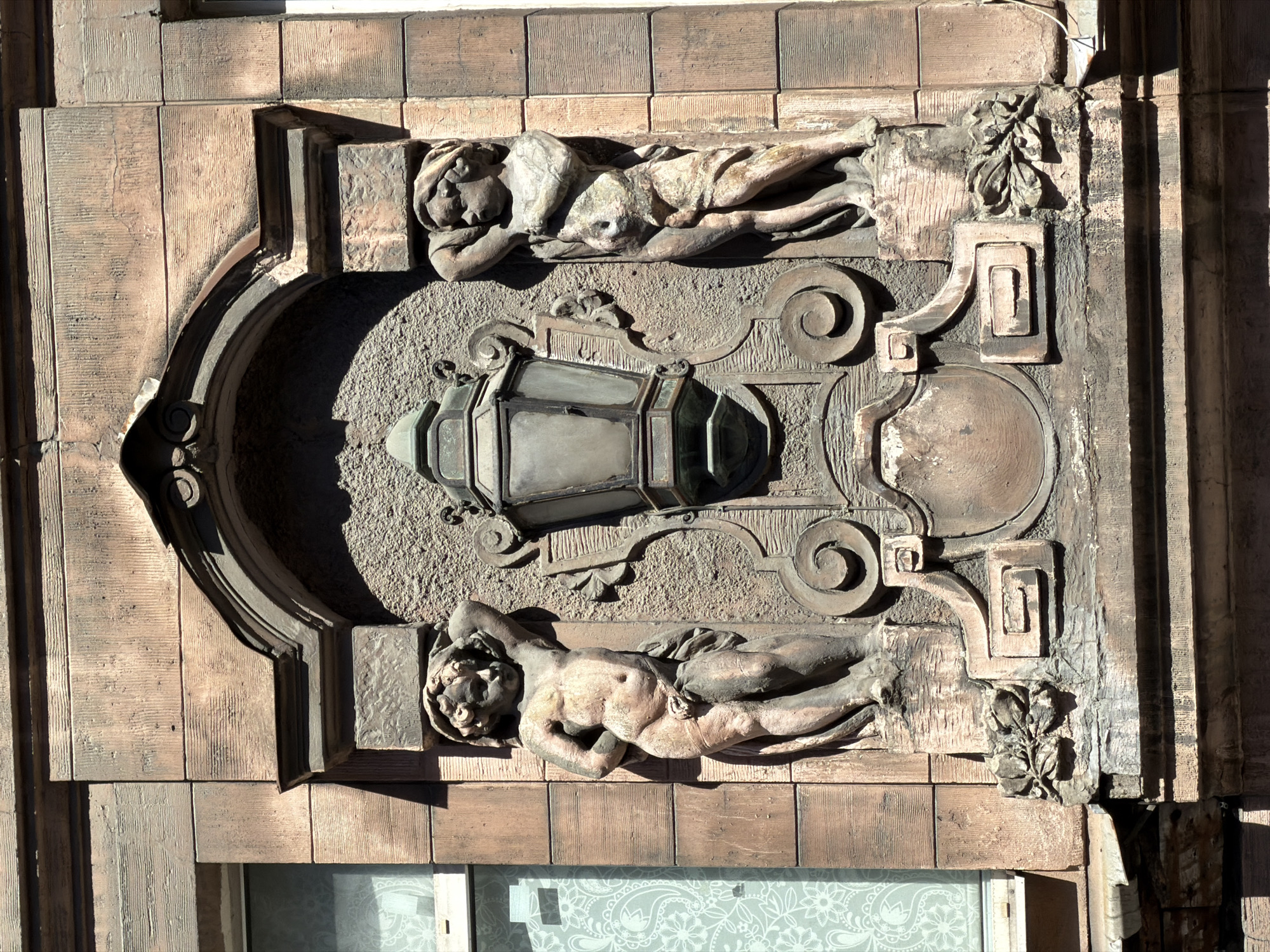 A detailed photograph captures a section of the facade of the Singer House (Dom Knigi / House of Books) at Nevsky Prospect, 28, Saint Petersburg, Russia. The scene is dominated by a central arched niche set within a light brown, horizontally striated stone or stucco facade.
Inside the niche, a large, ornate, dark-colored metal lantern with glass panes is mounted centrally. The lantern is supported by a decorative bracket featuring scrolled metalwork and stylized foliage. Flanking the lantern are two sculpted, nude male child figures, likely putti or cherubs, made of a lighter stone or concrete. The left figure stands with its left arm raised over its head and its right arm behind its back, a drapery covering its lower body. The right figure mirrors this pose, with its right arm raised and a similar drapery. Both figures exhibit signs of weathering and erosion.
Below the lantern and between the figures' pedestals, a decorative cartouche or shield-like element is visible, flanked by scrolls and floral or foliate motifs, resembling laurel wreaths. The niche itself is framed by classical architectural elements, including a curved architrave above.
To the left of the central ornamentation, a portion of a window with a white frame is visible, revealing a sheer white curtain adorned with a paisley-like floral pattern. A sliver of another window is visible on the far right. The facade material features a distinctive texture mimicking wood grain. The scene is illuminated by strong sunlight from the upper right, casting pronounced shadows, particularly on the left side of the niche and the left putto.