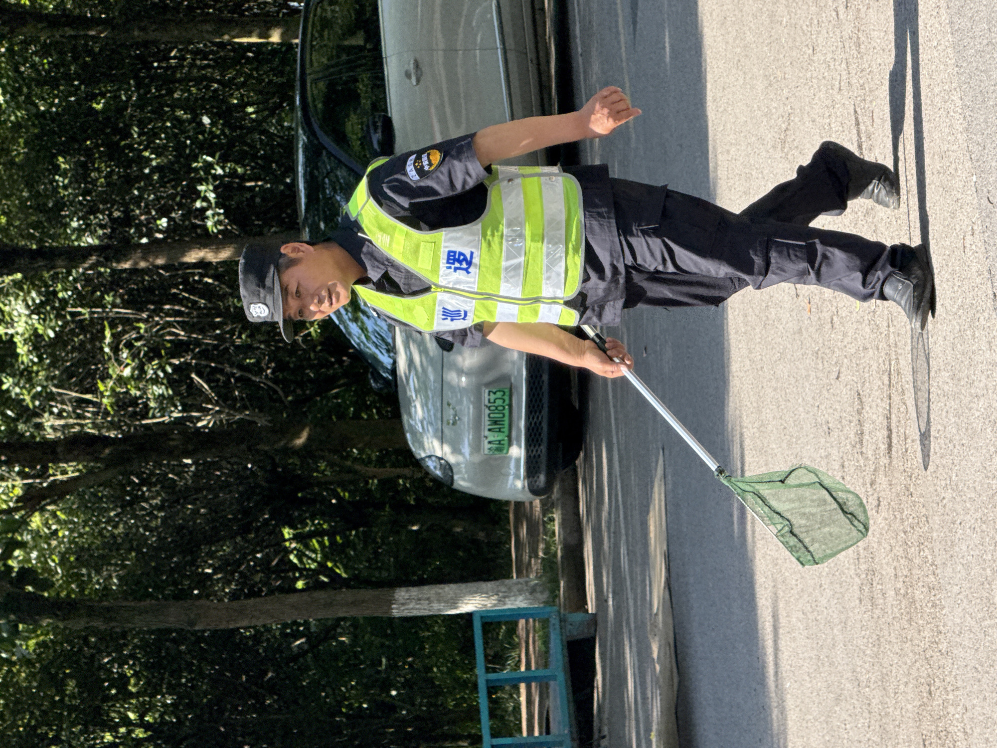 This image, captured in Jiangjin, China, depicts a man in uniform standing on a paved road under bright sunlight. He is dressed in a dark uniform, consisting of a long-sleeved shirt and trousers, topped with a neon yellow-green reflective vest featuring white stripes. A dark cap with an emblem is also part of his attire. The Chinese characters "巡逻" (Xúnluó), meaning "Patrol," are prominently displayed in blue on the front of his vest, identifying him as a patrol officer or security guard.
The man holds a long pole with a small green net attached to its end, poised as if in the midst of an activity or about to engage in one. He is looking towards the camera with a neutral expression. In the background, a light-colored car is parked, and its license plate, "川A AW0853," is clearly visible. The surrounding environment consists of dense green foliage and trees, suggesting a park, forest edge, or a treed street. Strong shadows are cast on the ground, indicating a clear, sunny day, likely during mid-morning or afternoon. A blue metal barrier or fence is partially visible in the lower left portion of the image.