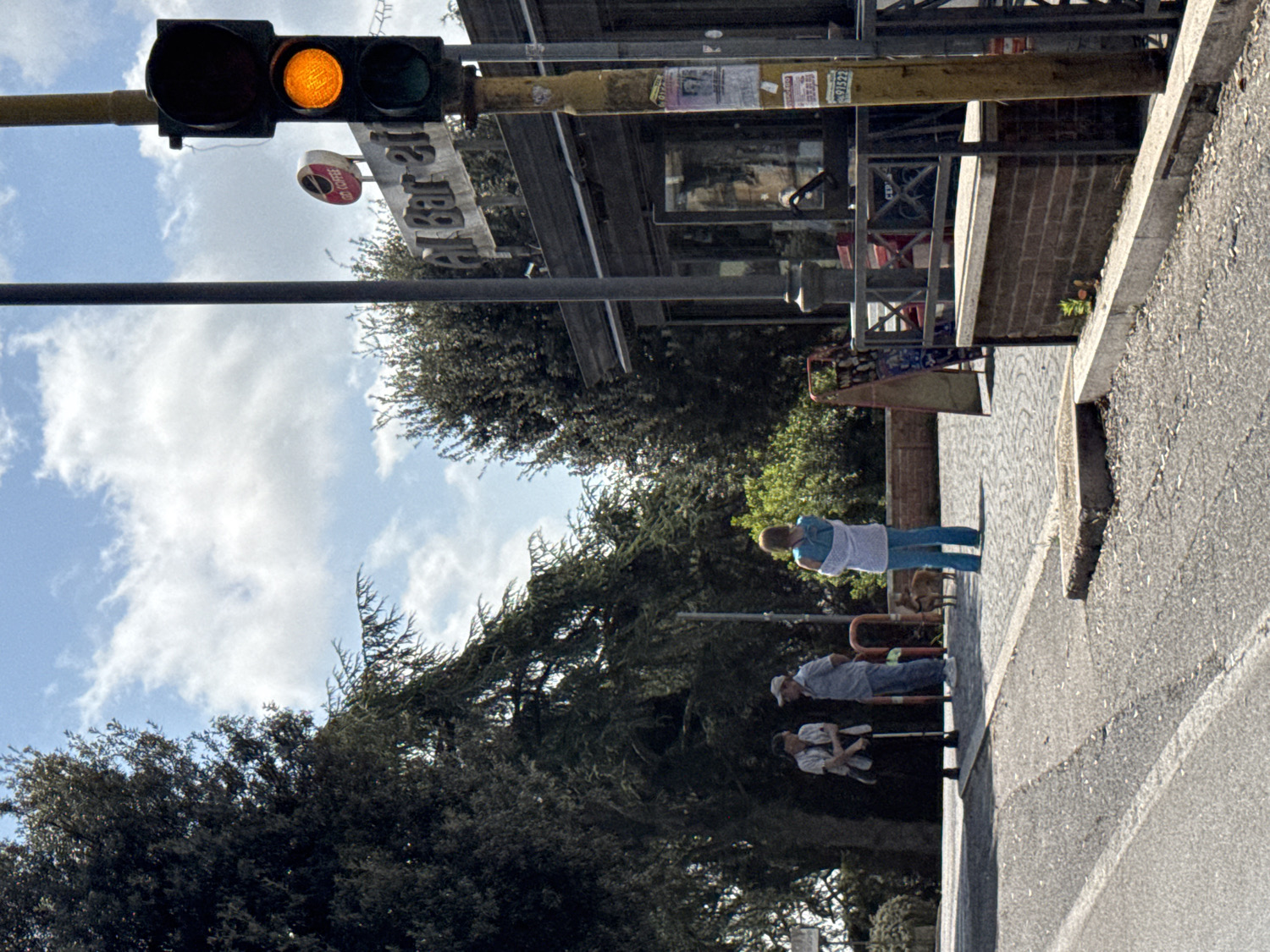 The image, oriented upright, captures a daytime street scene in Castel Gandolfo, Italy, under a clear sky with scattered white clouds. In the upper left, a traffic light displays a yellow (amber) signal. Below it, a vertical black pole runs down towards the bottom center.
On the right side of the image, a sidewalk runs parallel to an asphalt road, separated by a curb. Along the sidewalk, three individuals are present. Closest to the foreground, a woman wearing a light blue top and a white apron stands with her back to the viewer, looking slightly downwards. Near her feet, a small, light-colored animal, possibly a dog, is visible. Further along the sidewalk, a man in a light blue shirt and a white baseball cap stands. Next to him, another person, dressed in a white top and dark pants, is seated on a bench, facing away.
Behind the figures, mature trees with dense green foliage line the street. To the right of the traffic light and above the sidewalk, a building facade with dark windows and brickwork is visible, possibly part of a commercial establishment. A sign bearing the inverted text "Bar 21" hangs from an awning or structure above the building entrance. Another circular sign, also inverted, reads "COFFEE". Yellow horizontal beams running across the top of the building display some unreadable posters or notices. The overall impression is a calm, sunny day in a European town.