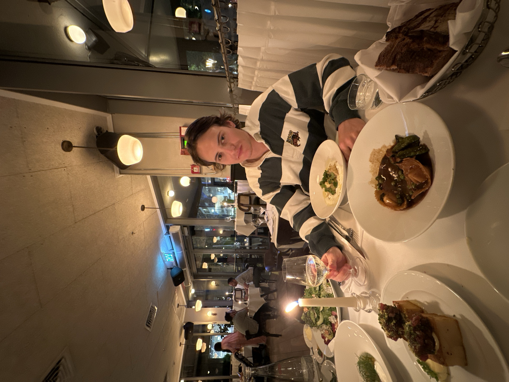 The image captures an indoor scene at a restaurant in Tel-Aviv, Israel, likely during the evening due to the dim, warm artificial lighting. The perspective is slightly angled, showing a person seated at a white-tablecloth-covered table in the foreground, with other diners and restaurant decor in the background.
The main subject, a person with short, dark hair, wears a striped long-sleeve rugby shirt in green/teal and white, featuring a "BISON" logo with a buffalo emblem on the left chest. They are looking directly at the camera with a neutral expression. Their right hand is visible near a wine glass on the table.
The table setting includes white ceramic plates with various dishes. In front of the main subject are a small bowl with a light-colored, herb-garnished dish and a larger plate holding a saucy main course, possibly meat or fish, accompanied by green vegetables and grains. Other visible dishes on the table include a basket of dark bread, a salad, and a plate with what appears to be golden-brown squares topped with a dark relish. A tall white candle in a clear holder provides additional light.
In the background, several other patrons are visible, seated at their own tables, engaged in dining or conversation. The restaurant features modern, glowing circular and oval light fixtures on the ceiling and walls. Large windows separate the dining area from an outdoor or adjacent space, revealing more distant lights. A blue sign displaying the Hebrew word "יציאה" (Yetsia), meaning "EXIT," is clearly visible above the windows, confirming the Israeli location. Air conditioning vents and what appear to be red fire alarm components are also present on the walls. The overall ambiance is relaxed and sophisticated.