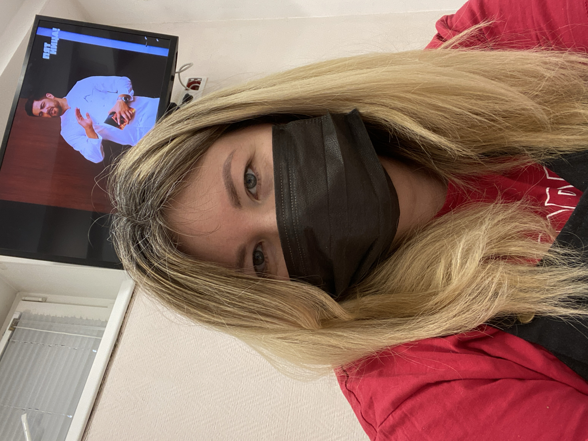 A woman wearing a black face mask and a red shirt is lying down. Her blonde hair is spread around her face. Behind her is a TV showing a man in a white shirt holding an object. The scene appears to be indoors, with a window visible to the left of the TV. The location is Chaikovsky, Russia.